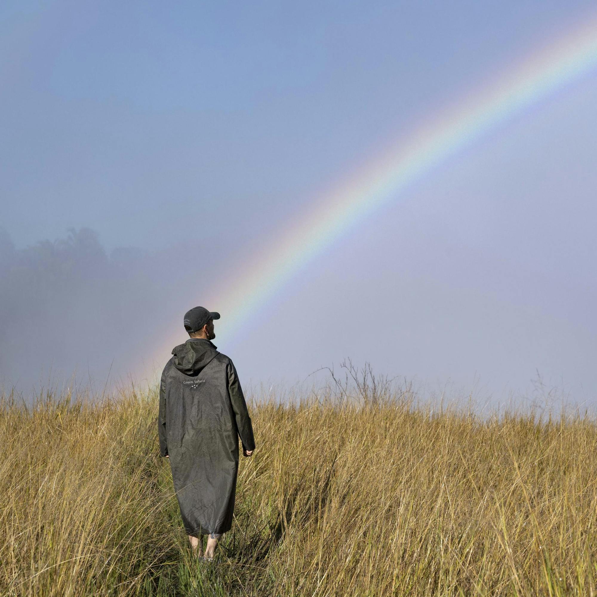 A traveler stands in tall golden grass as a bright rainbow arcs across a misty blue sky after the rain nearby.