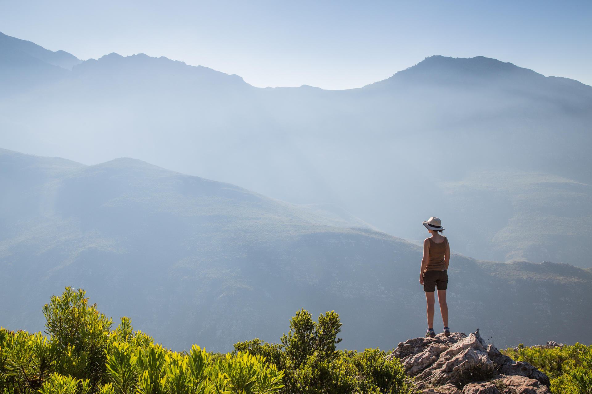 A hiker stands on a rocky viewpoint overlooking layered blue mountains and valleys fading into morning haze.