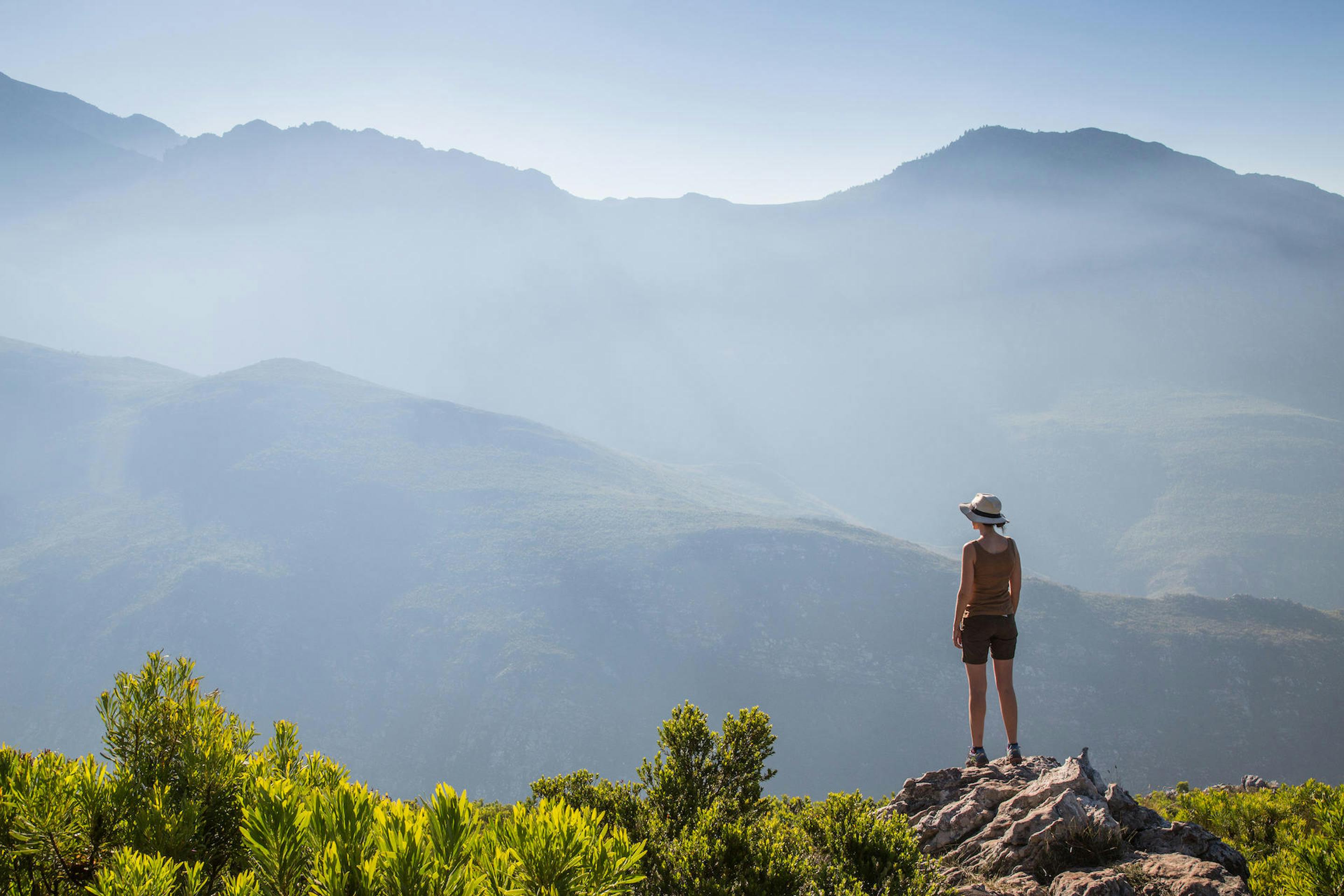 A hiker stands on a rocky viewpoint overlooking layered blue mountains and valleys fading into morning haze.