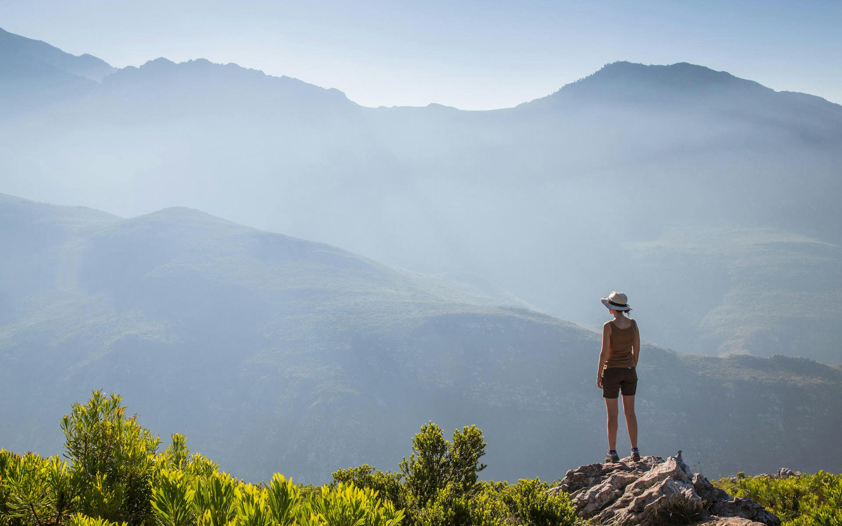 A hiker stands on a rocky viewpoint overlooking layered blue mountains and valleys fading into morning haze.