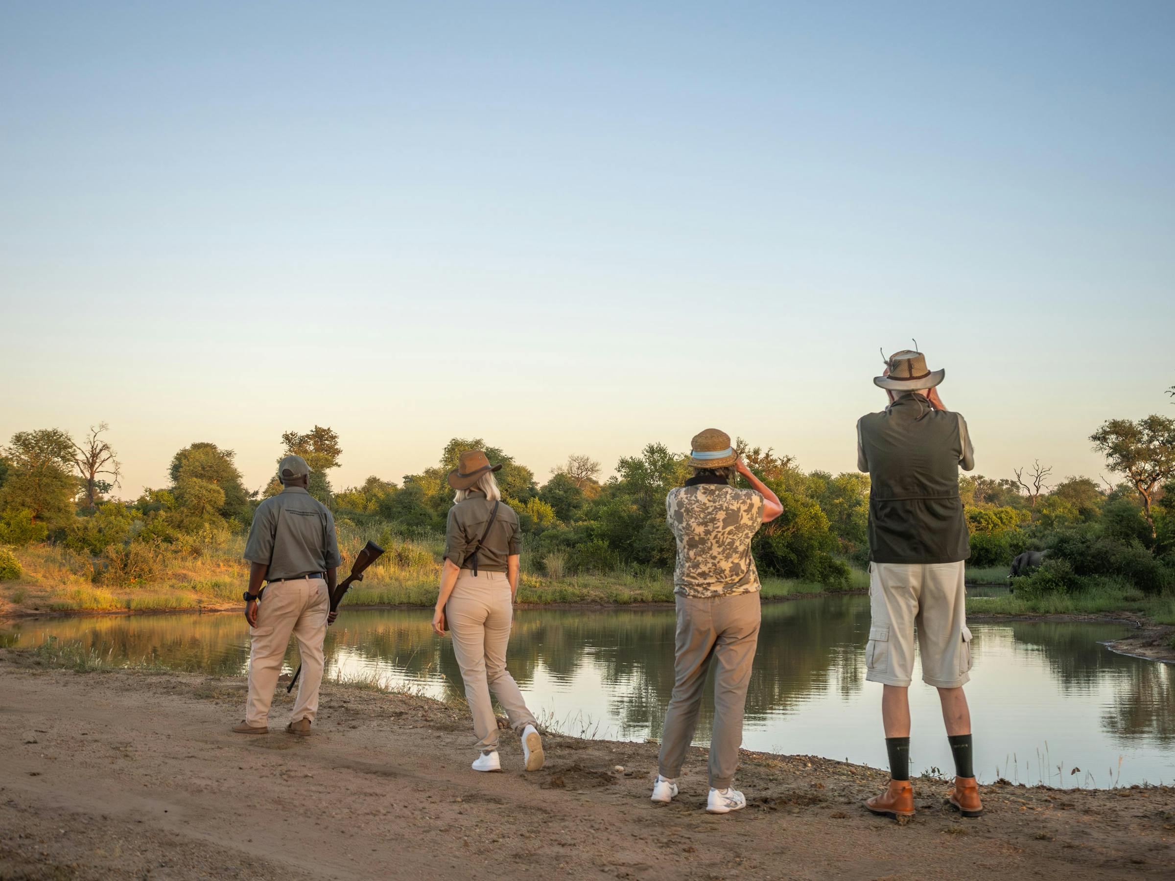 Four people pause on a riverside track at sunset, looking across still water toward distant trees and reeds.