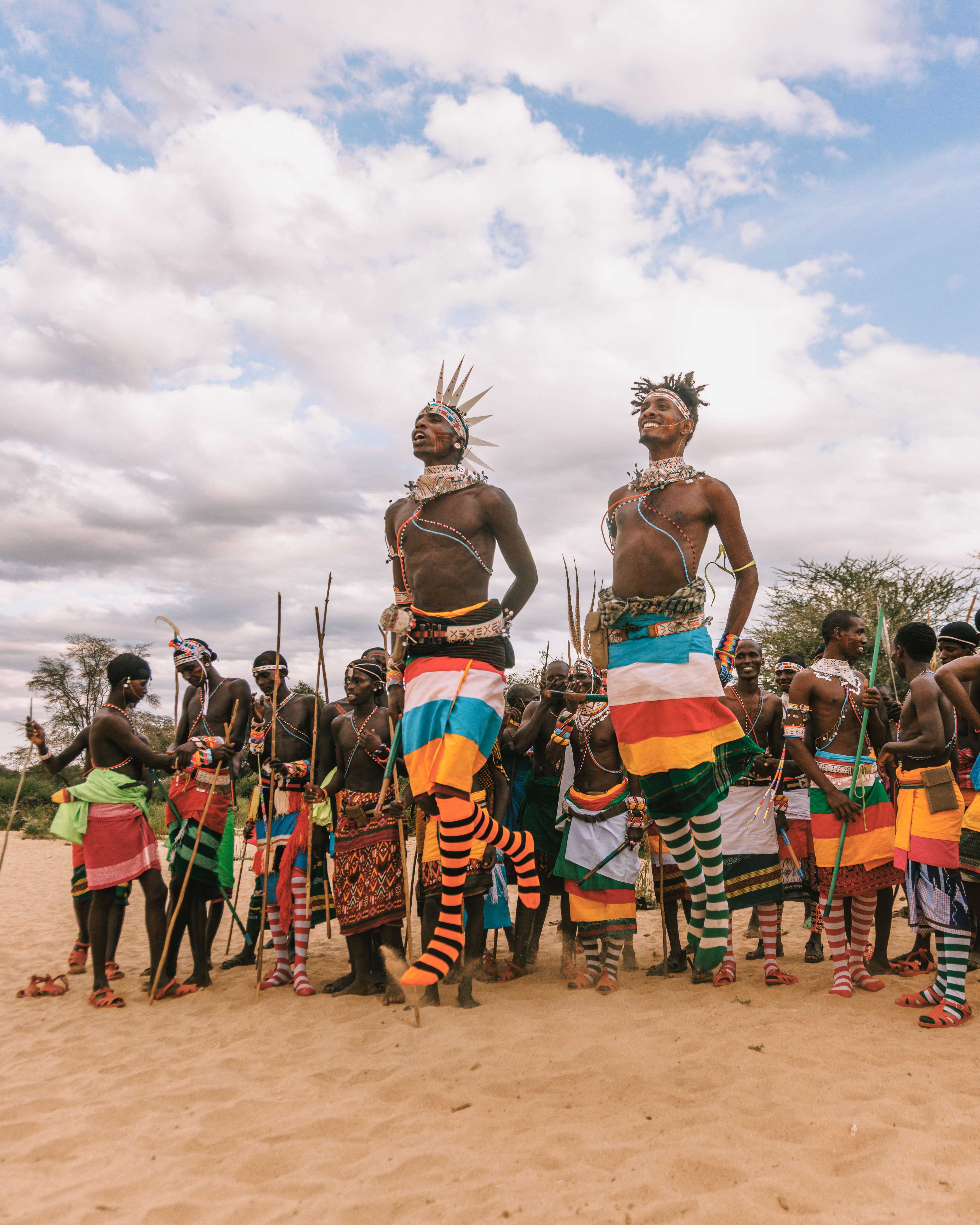 Samburu dancers in bright beaded regalia perform on sandy ground as a crowd watches under a wide cloudy sky.