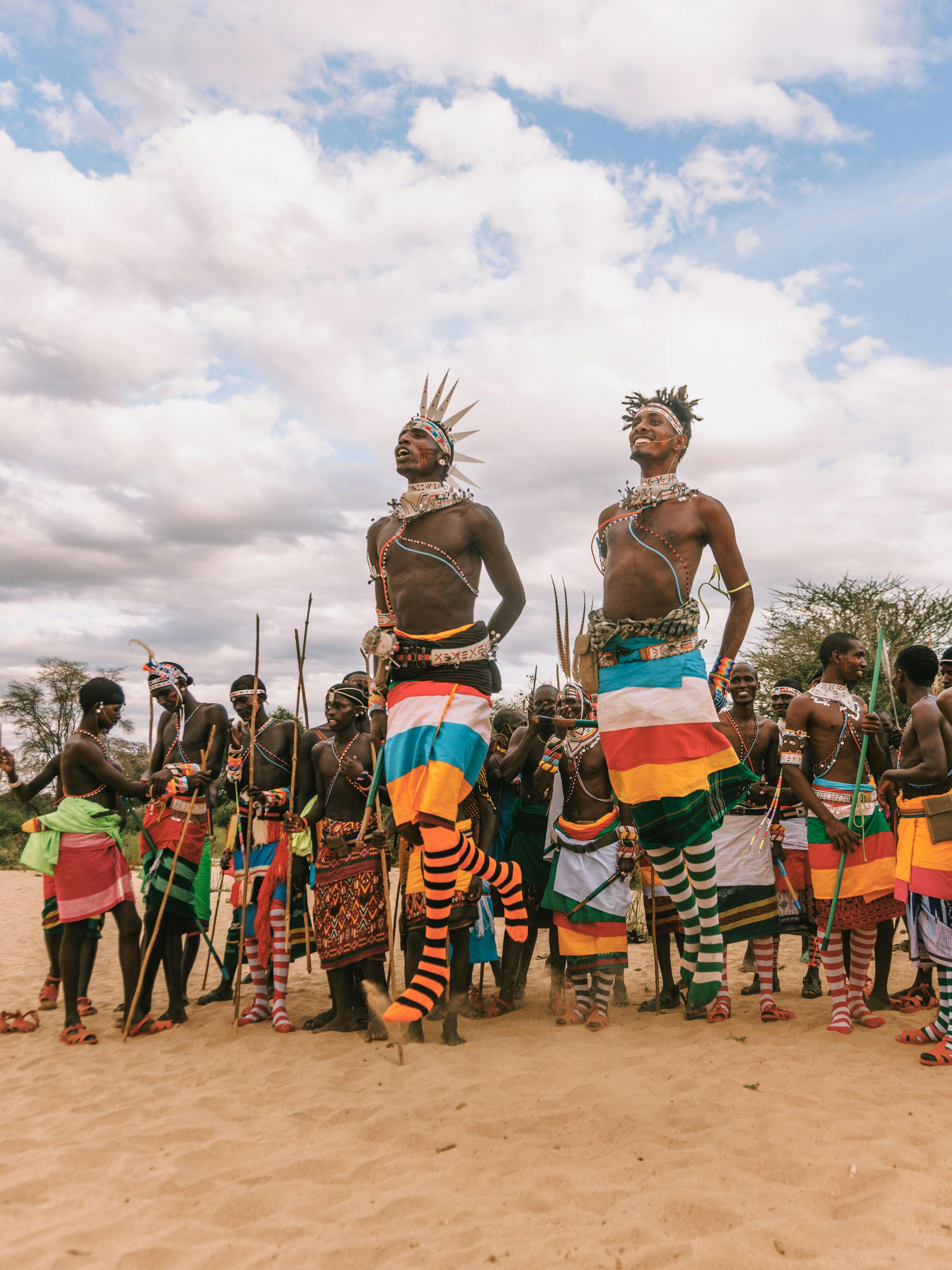 Samburu dancers in bright beaded regalia perform on sandy ground as a crowd watches under a wide cloudy sky.