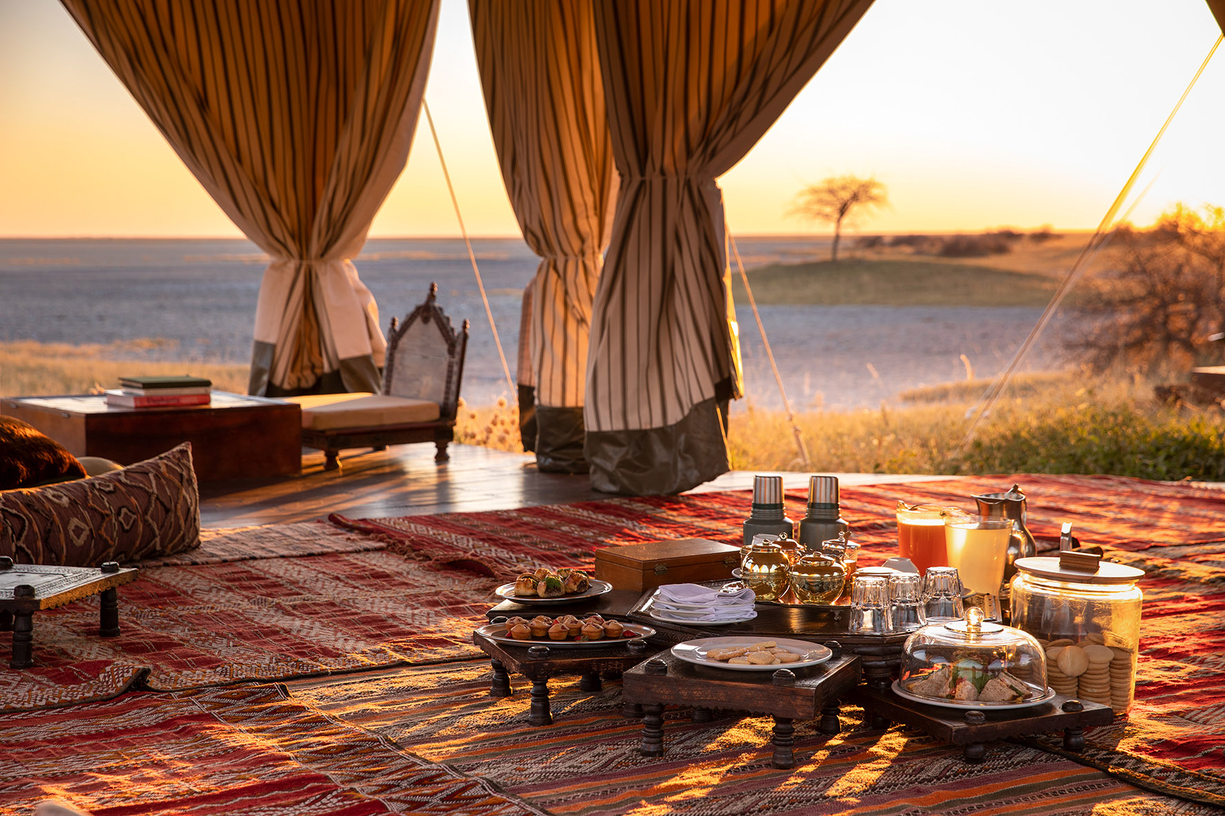 A low table set for tea sits inside a billowing canvas tent, with patterned rugs and an open horizon view.