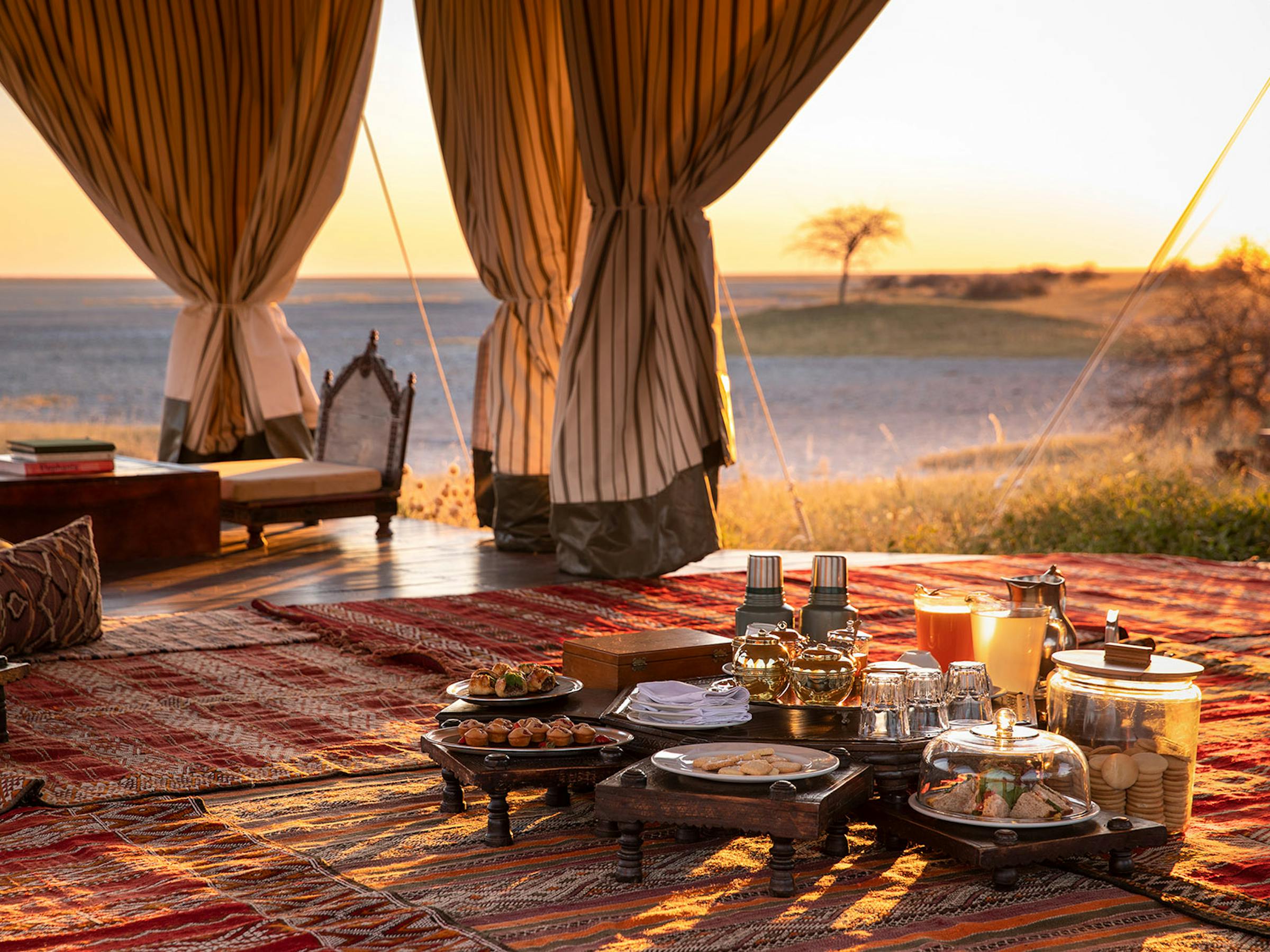 A low table set for tea sits inside a billowing canvas tent, with patterned rugs and an open horizon view.