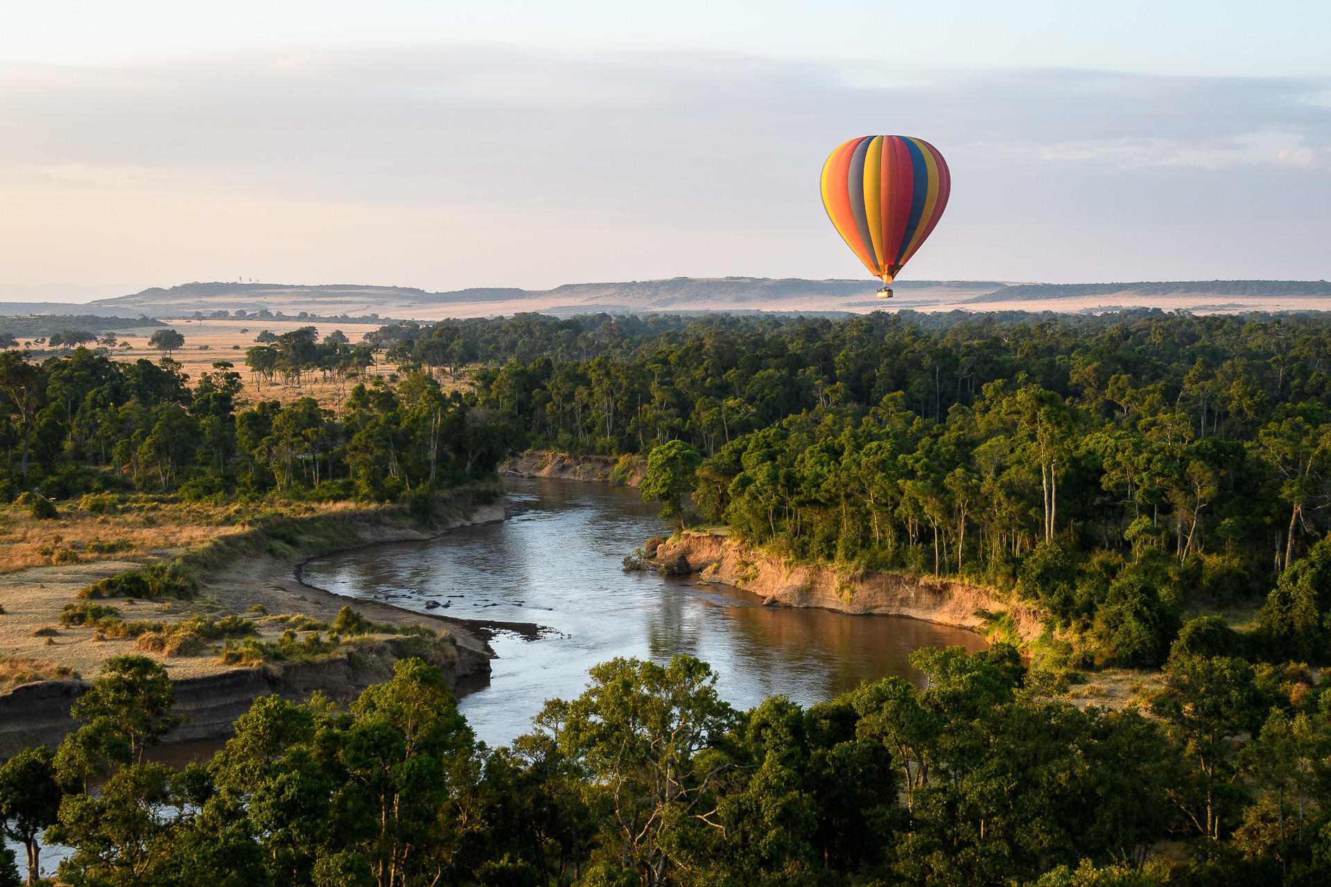 Hot-air balloon floats above a winding river and green forest, with open savanna stretching to the horizon.