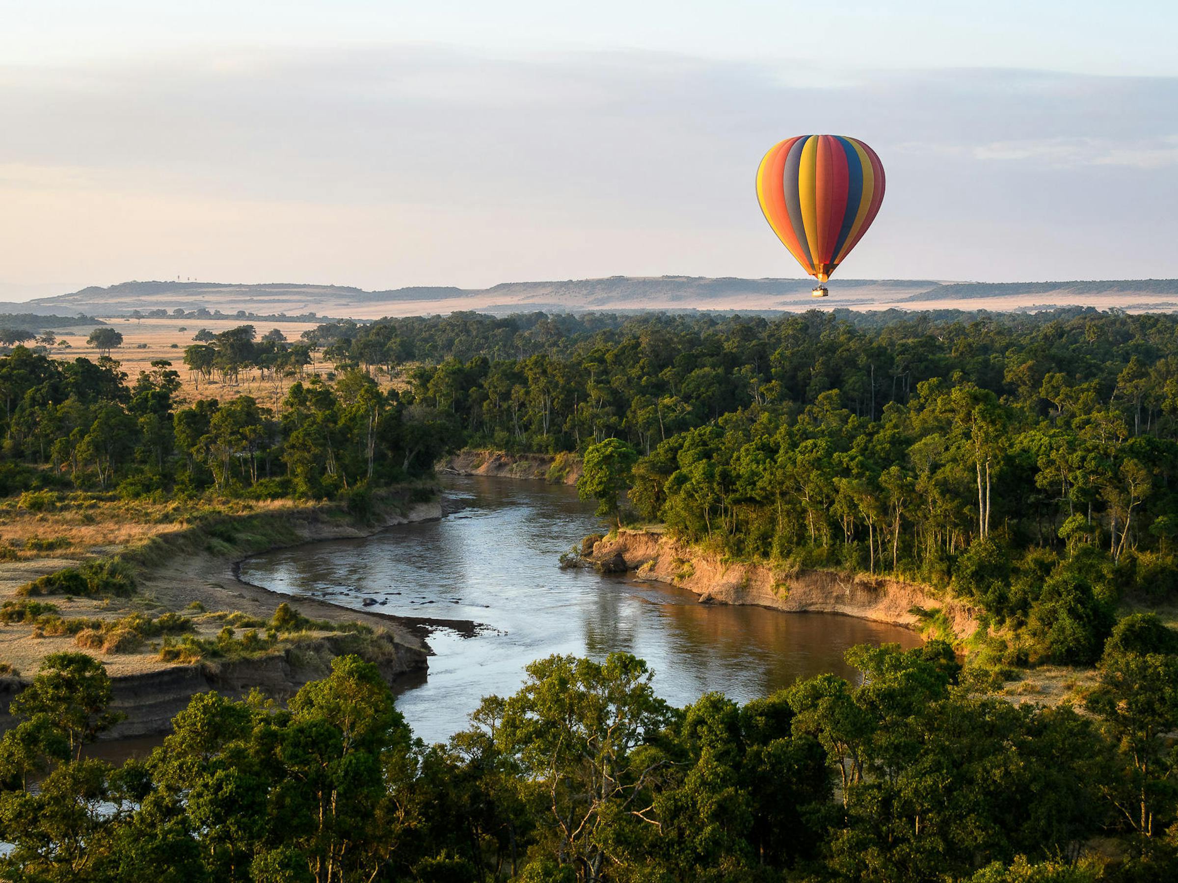 Hot-air balloon floats above a winding river and green forest, with open savanna stretching to the horizon.