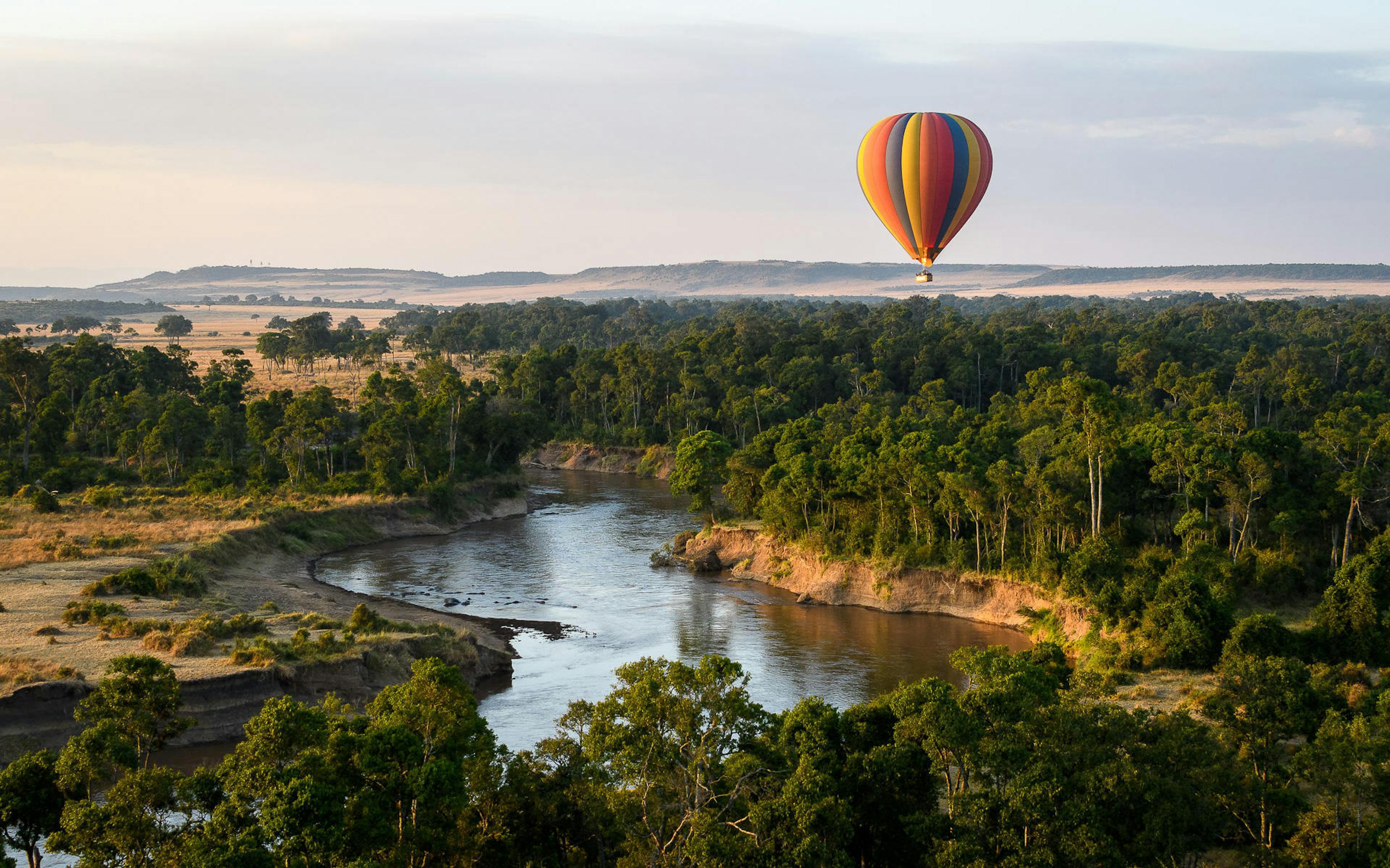 Hot-air balloon floats above a winding river and green forest, with open savanna stretching to the horizon.