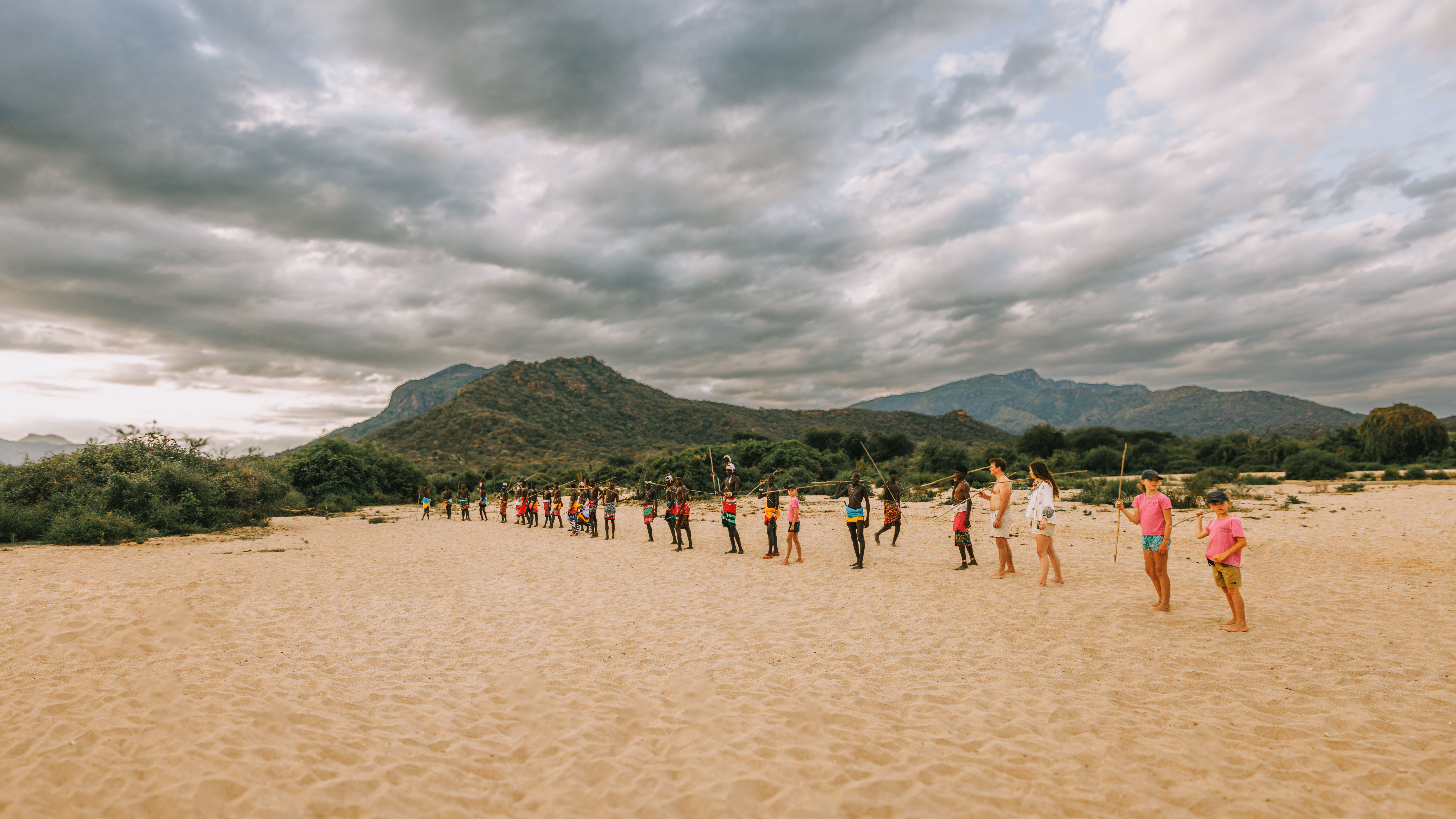 A line of people walks across a sandy plain toward a distant hill, beneath dramatic gray clouds overhead.