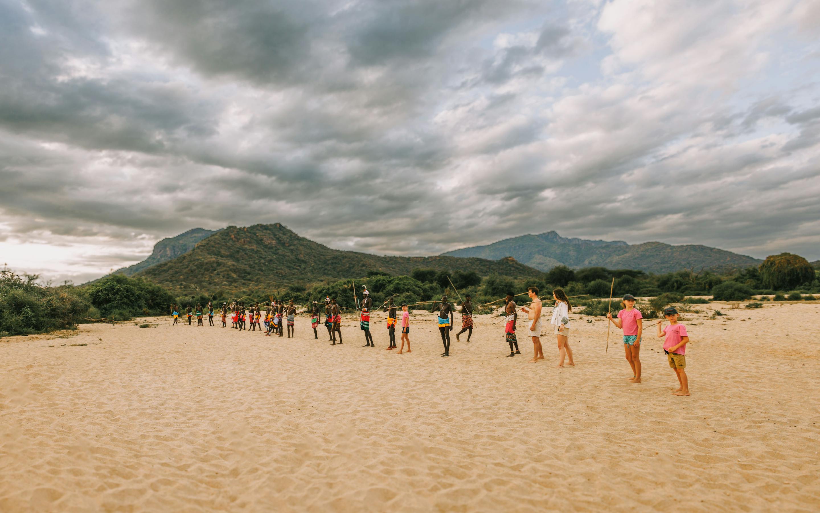 A line of people walks across a sandy plain toward a distant hill, beneath dramatic gray clouds overhead.