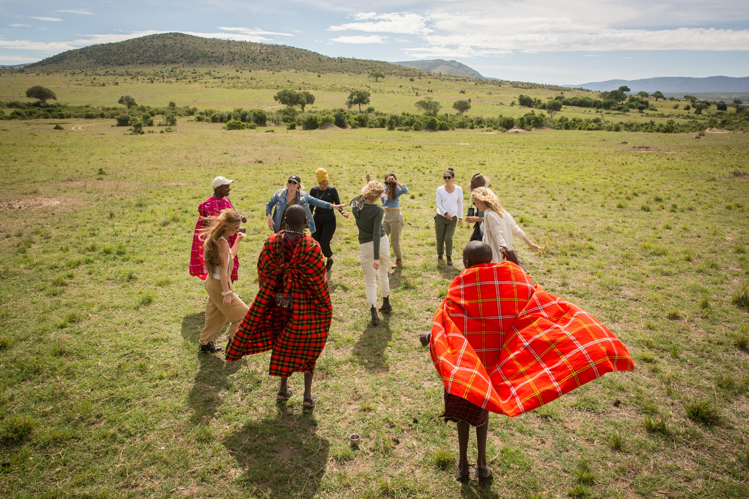Guests and Maasai guides walk across open savannah grassland, red shukas bright against a pale sky beyond.