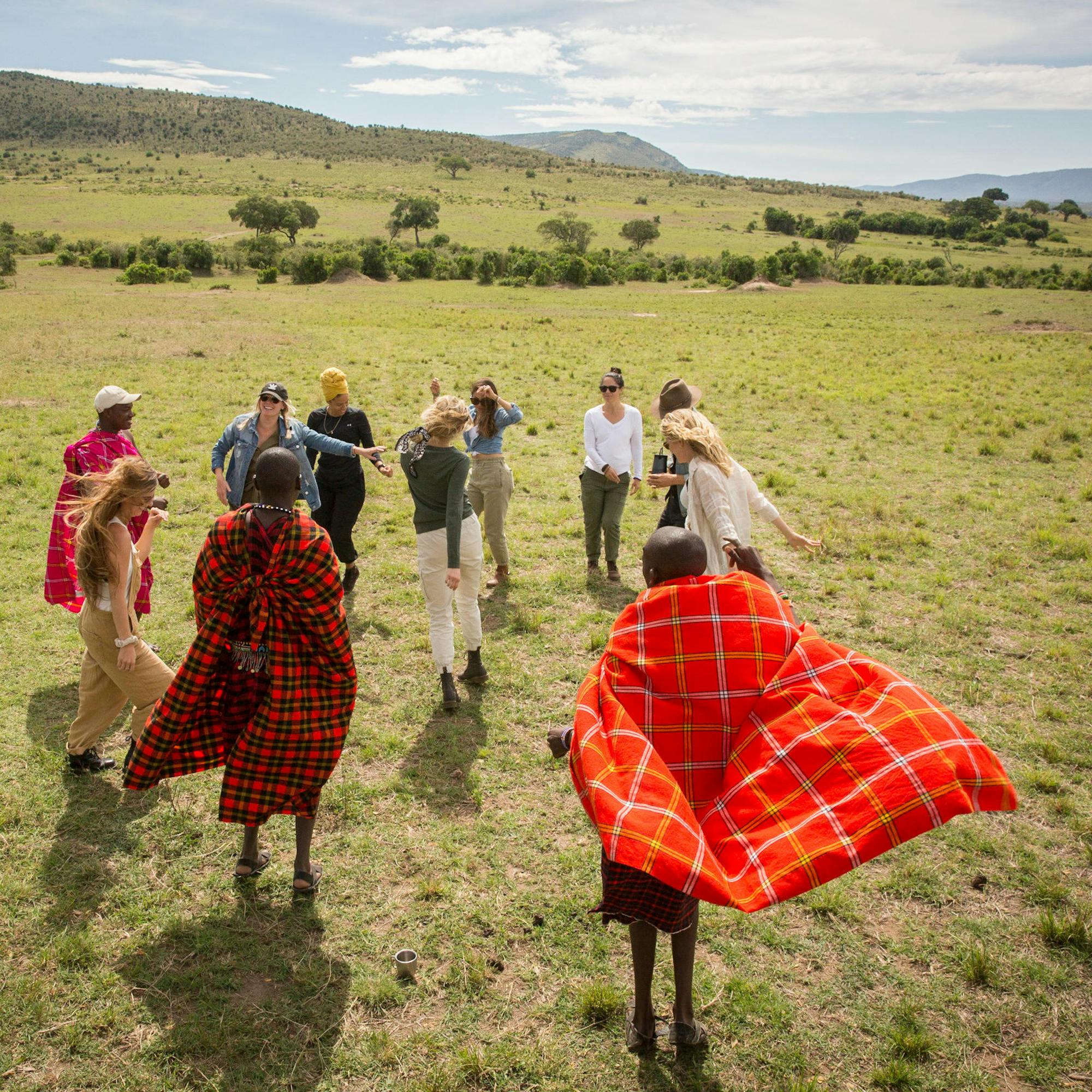 Guests and Maasai guides walk across open savannah grassland, red shukas bright against a pale sky beyond.