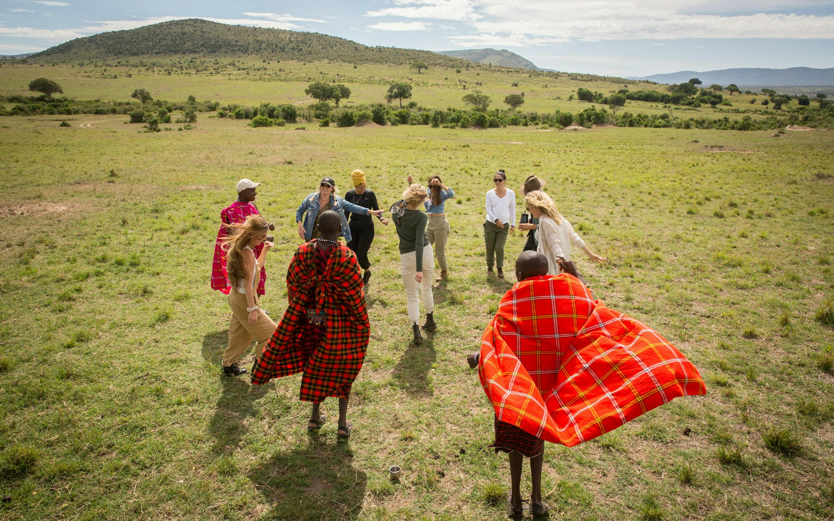 Guests and Maasai guides walk across open savannah grassland, red shukas bright against a pale sky beyond.
