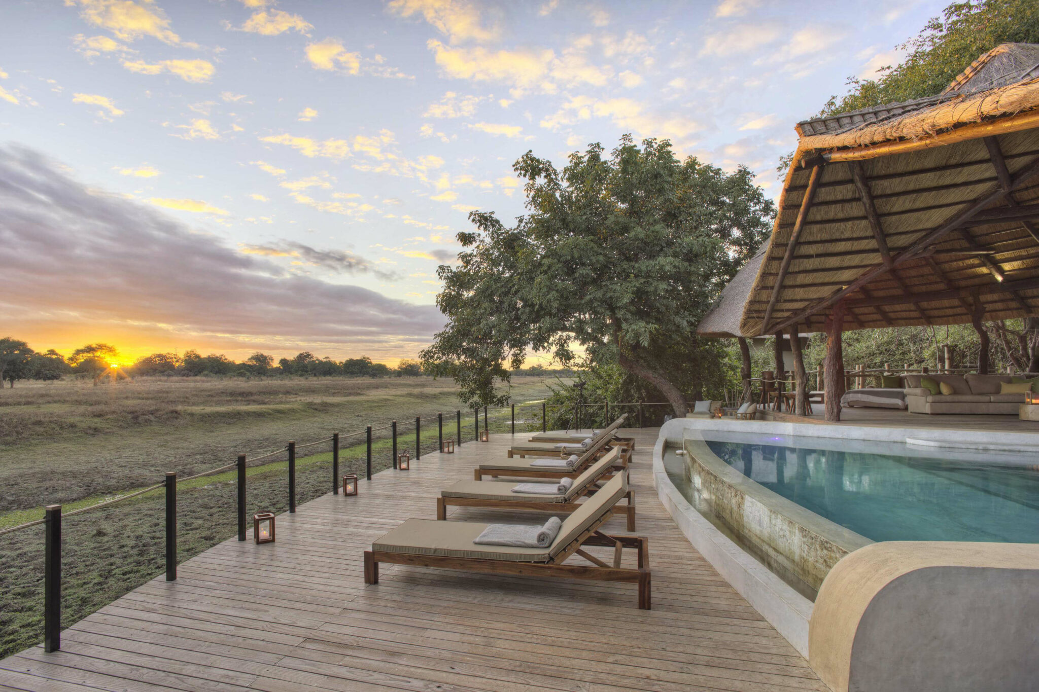 Sun loungers line a pool beside an open-sided lodge, looking out to wide plains as the sun sets behind clouds.