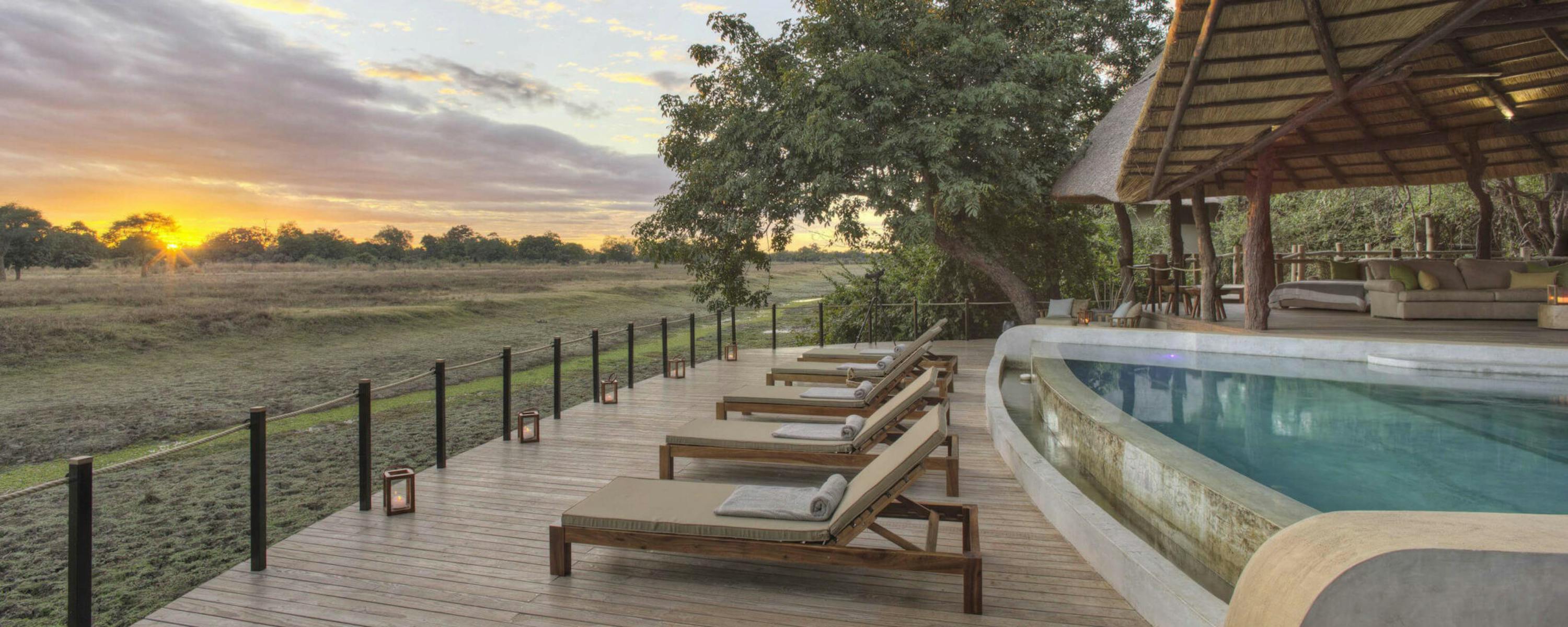 Sun loungers line a pool beside an open-sided lodge, looking out to wide plains as the sun sets behind clouds.