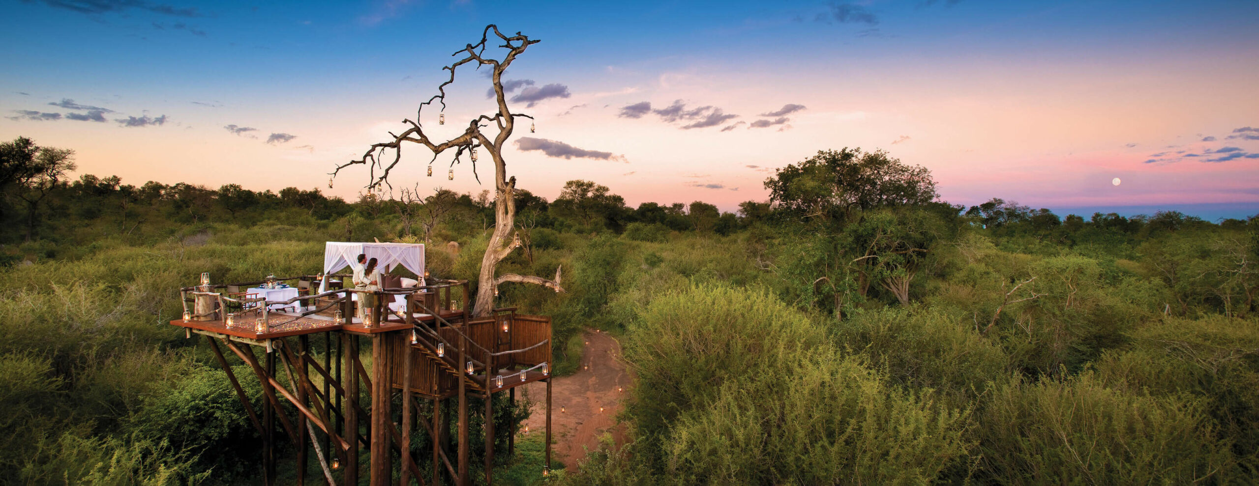 An elevated dining deck wraps around a bare tree at dusk, with a set table overlooking green bush and pink sky.