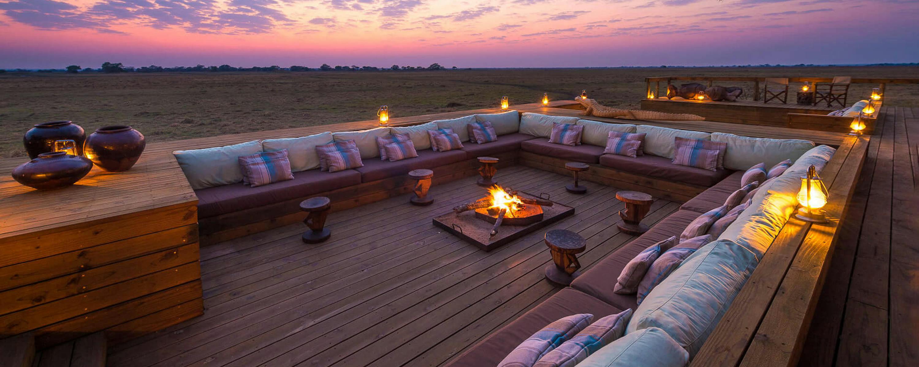 A cushioned lounge deck glows with lanterns at sunset, facing open plains under a dramatic purple sky and clouds.