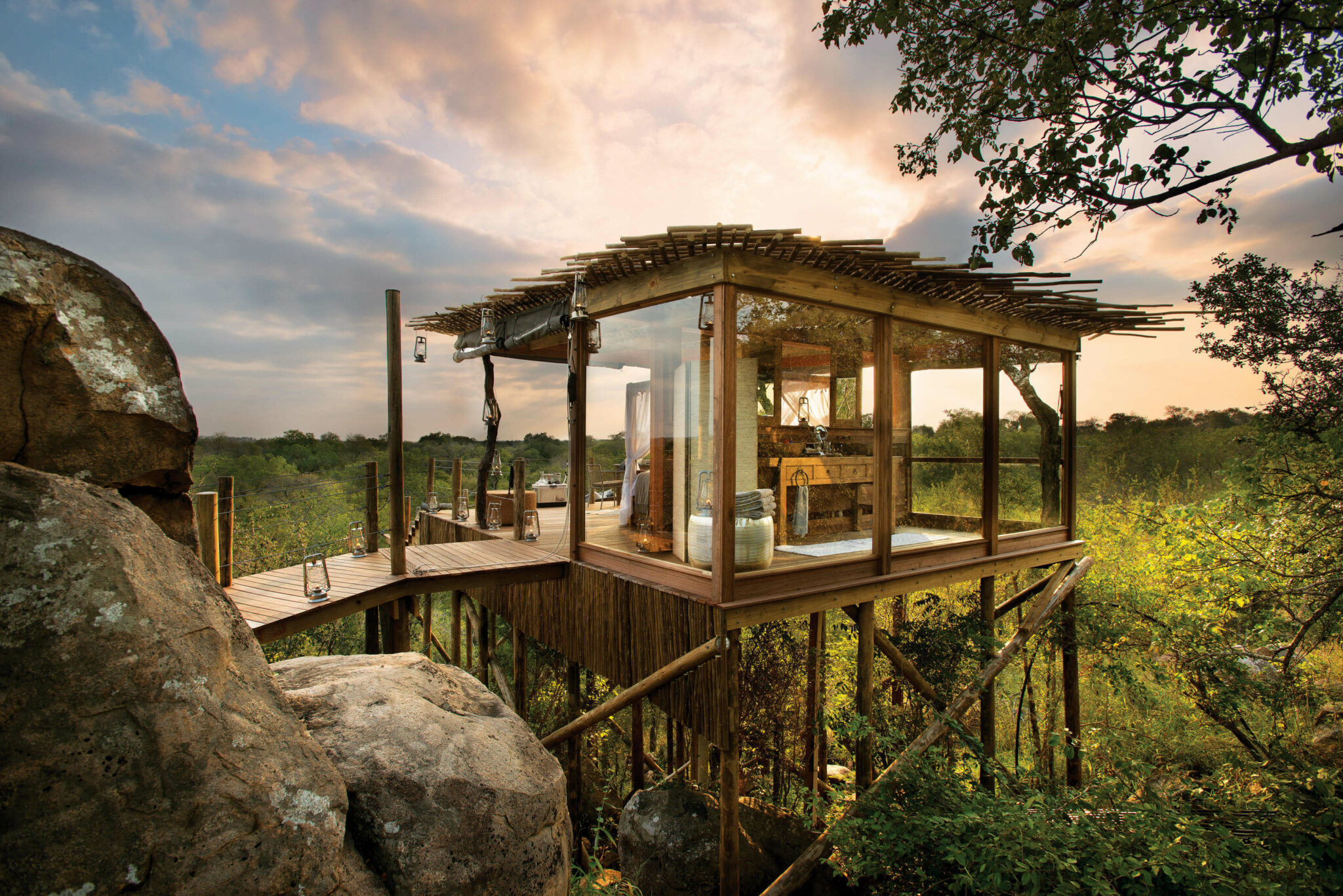 A glass-walled treehouse suite stands on stilts above rocks, surrounded by green savanna in warm evening light.