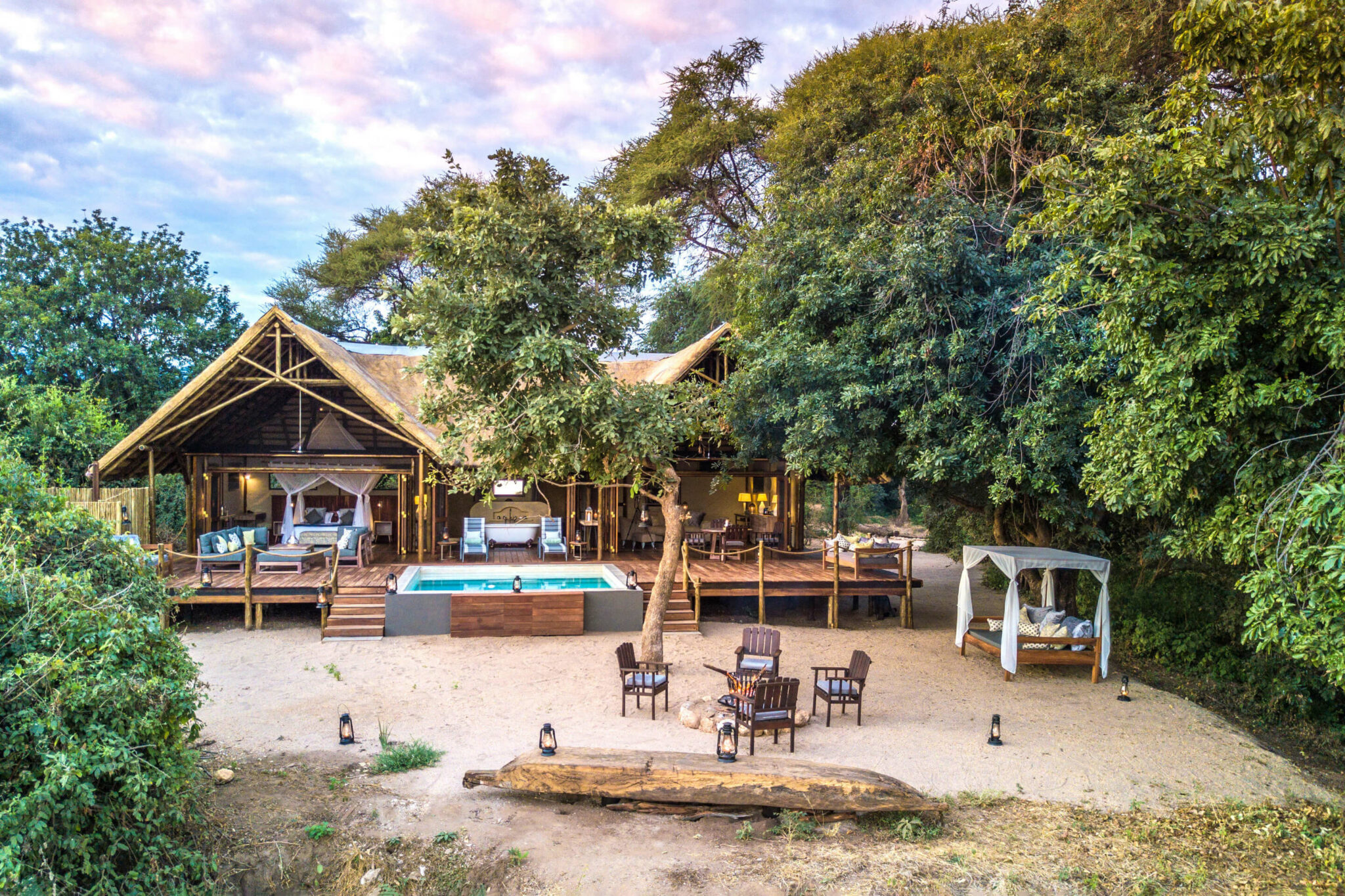 A thatched lodge and pool sit in a sandy clearing, with tables set under trees and a soft pastel sky overhead.