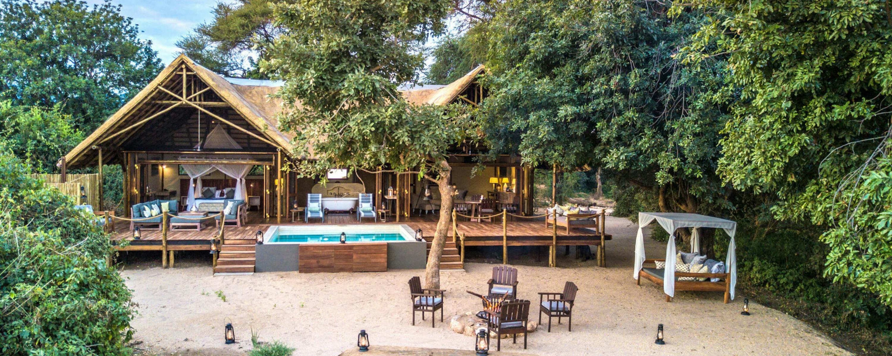 A thatched lodge and pool sit in a sandy clearing, with tables set under trees and a soft pastel sky overhead.