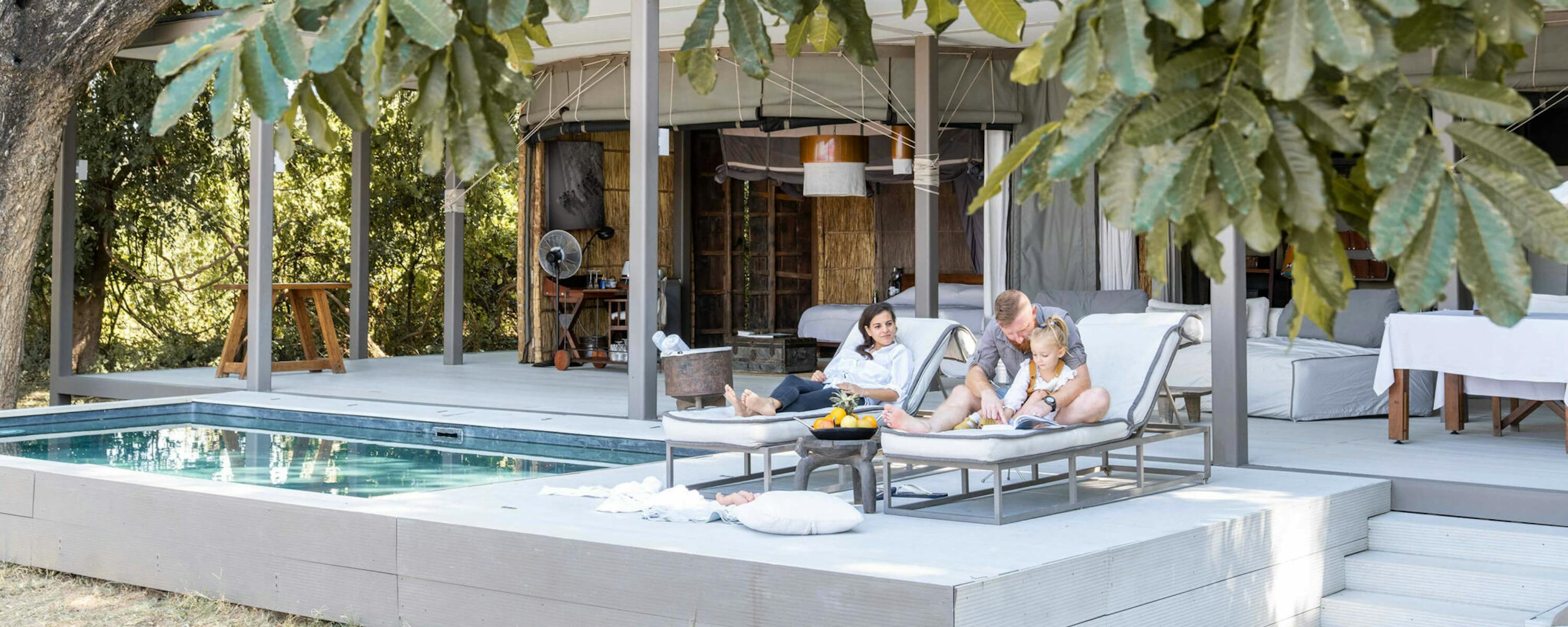 Guests relax on a shaded terrace beside a long pool at a modern lodge, with seating under broad green leaves.