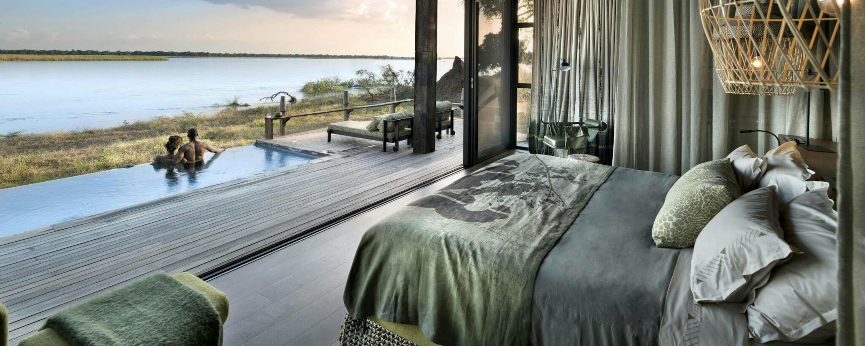 Woven pendant lamps and a netted swing frame a river view from a shaded veranda, with reeds and islands below.