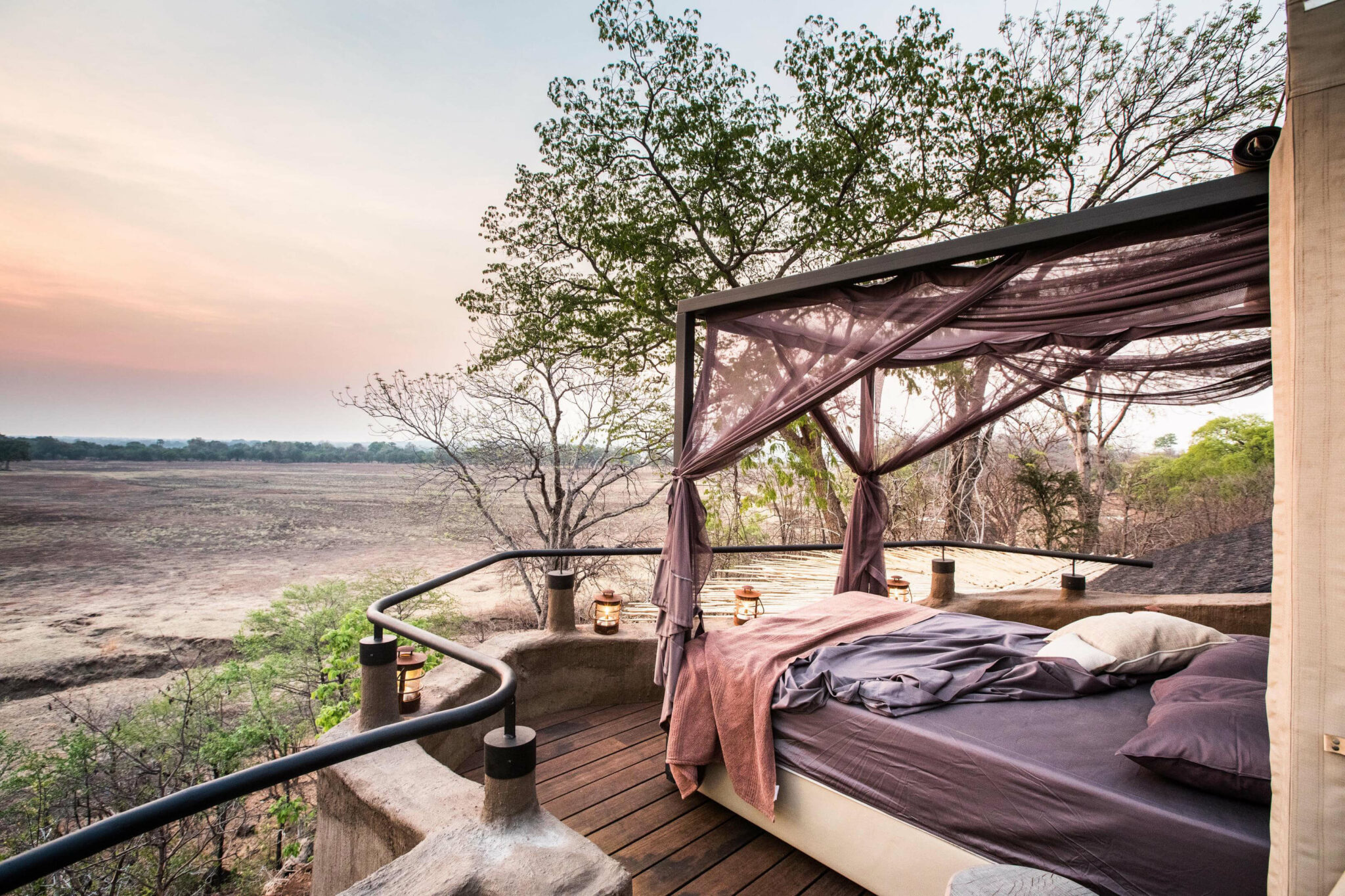 A canopy bed sits on a wooden deck overlooking a dry riverbed, with trees and pink twilight on the horizon.