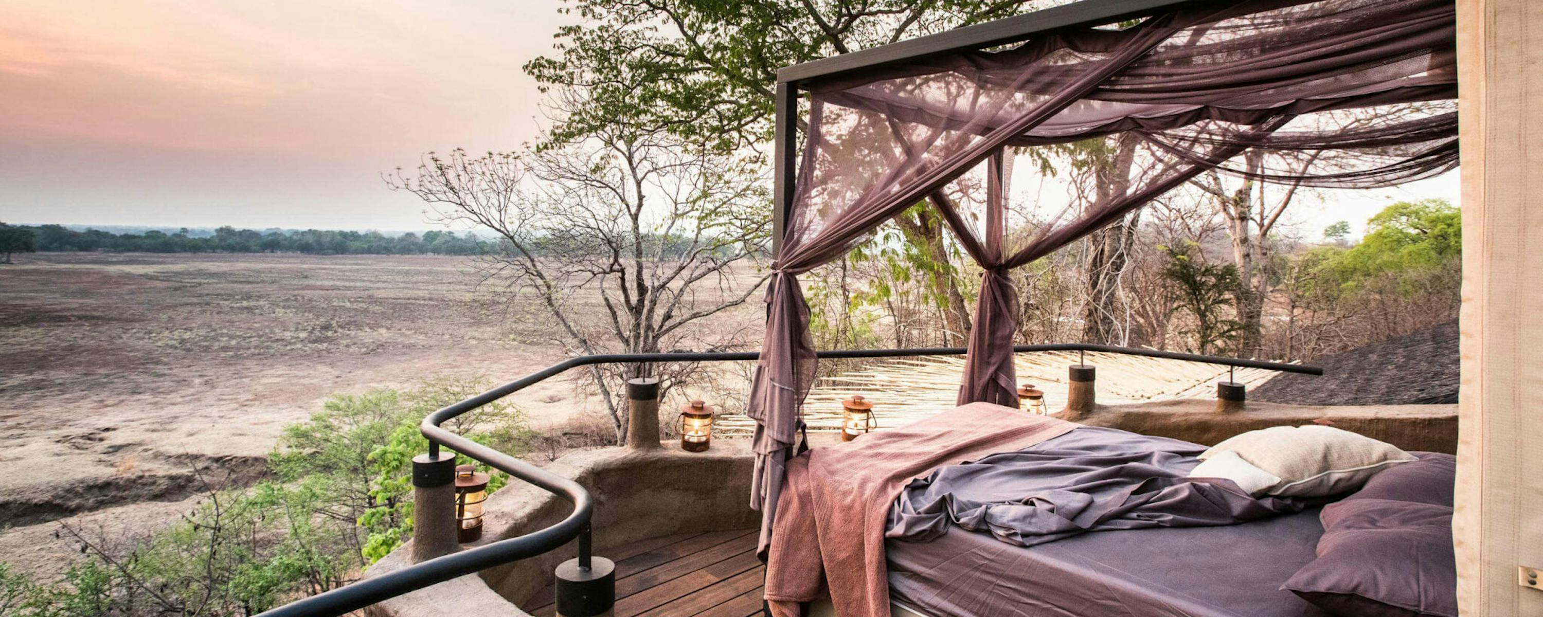 A canopy bed sits on a wooden deck overlooking a dry riverbed, with trees and pink twilight on the horizon.