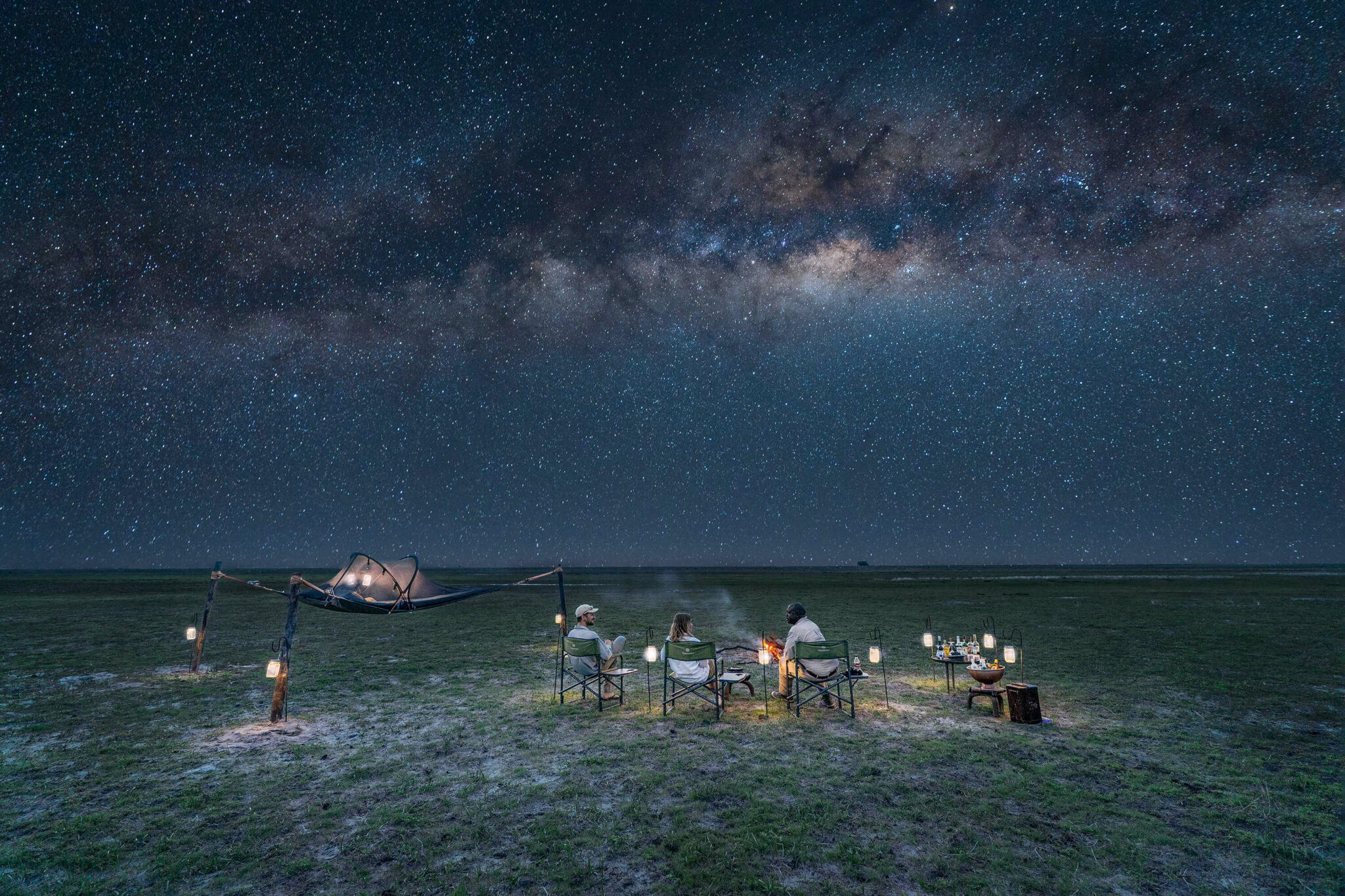 Lanterns and camp chairs sit on open grass beneath the Milky Way, with a small tent glowing in the distance.