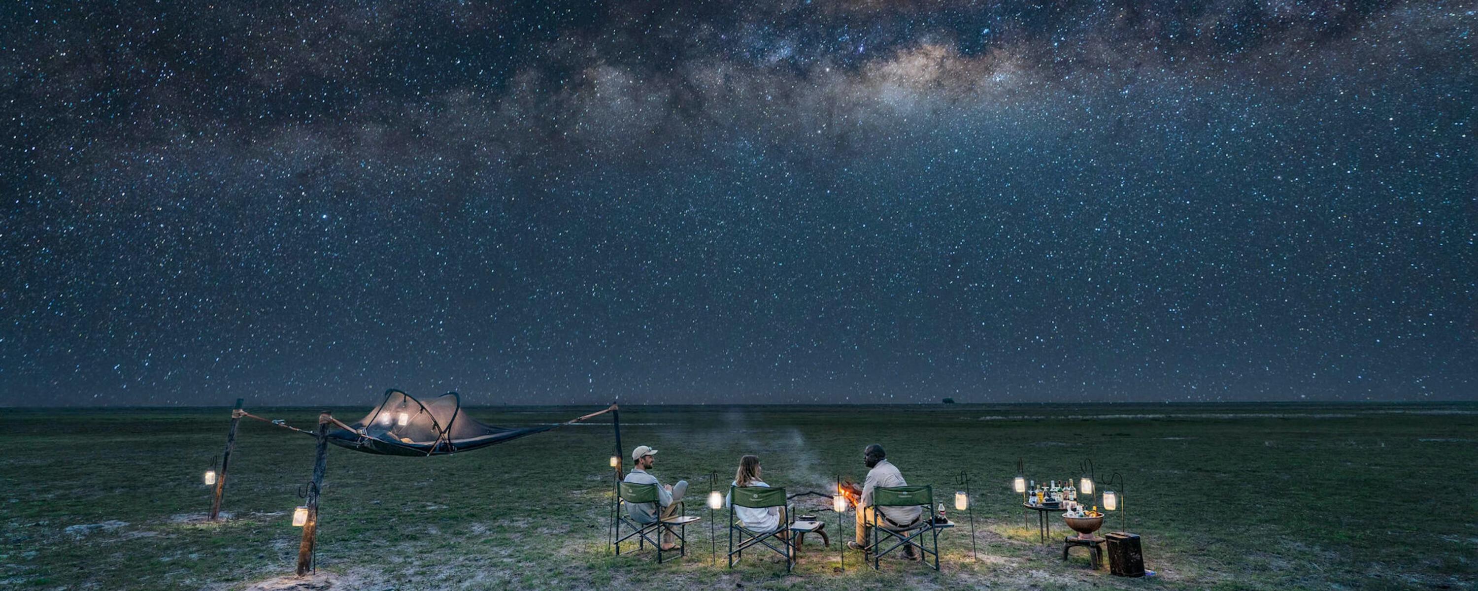 Lanterns and camp chairs sit on open grass beneath the Milky Way, with a small tent glowing in the distance.
