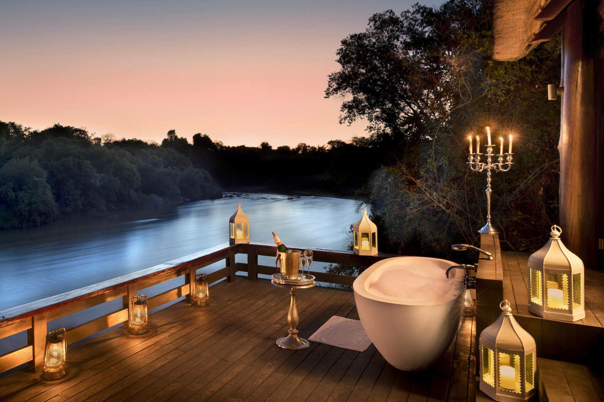 A freestanding bathtub sits on a riverside deck at dusk, lit by lanterns and candles under a soft pink sky.