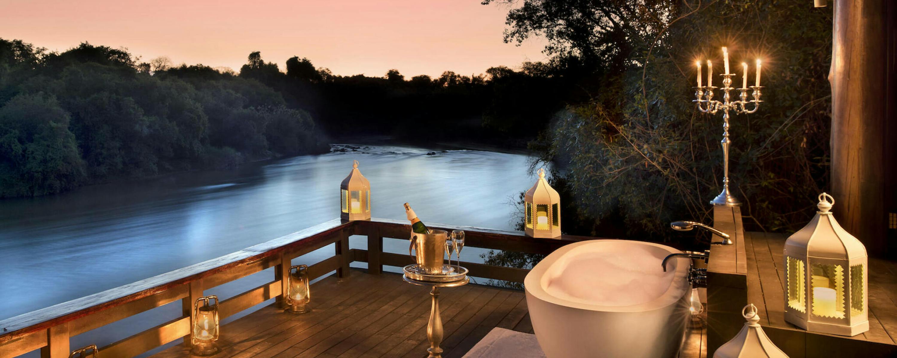 A freestanding bathtub sits on a riverside deck at dusk, lit by lanterns and candles under a soft pink sky.