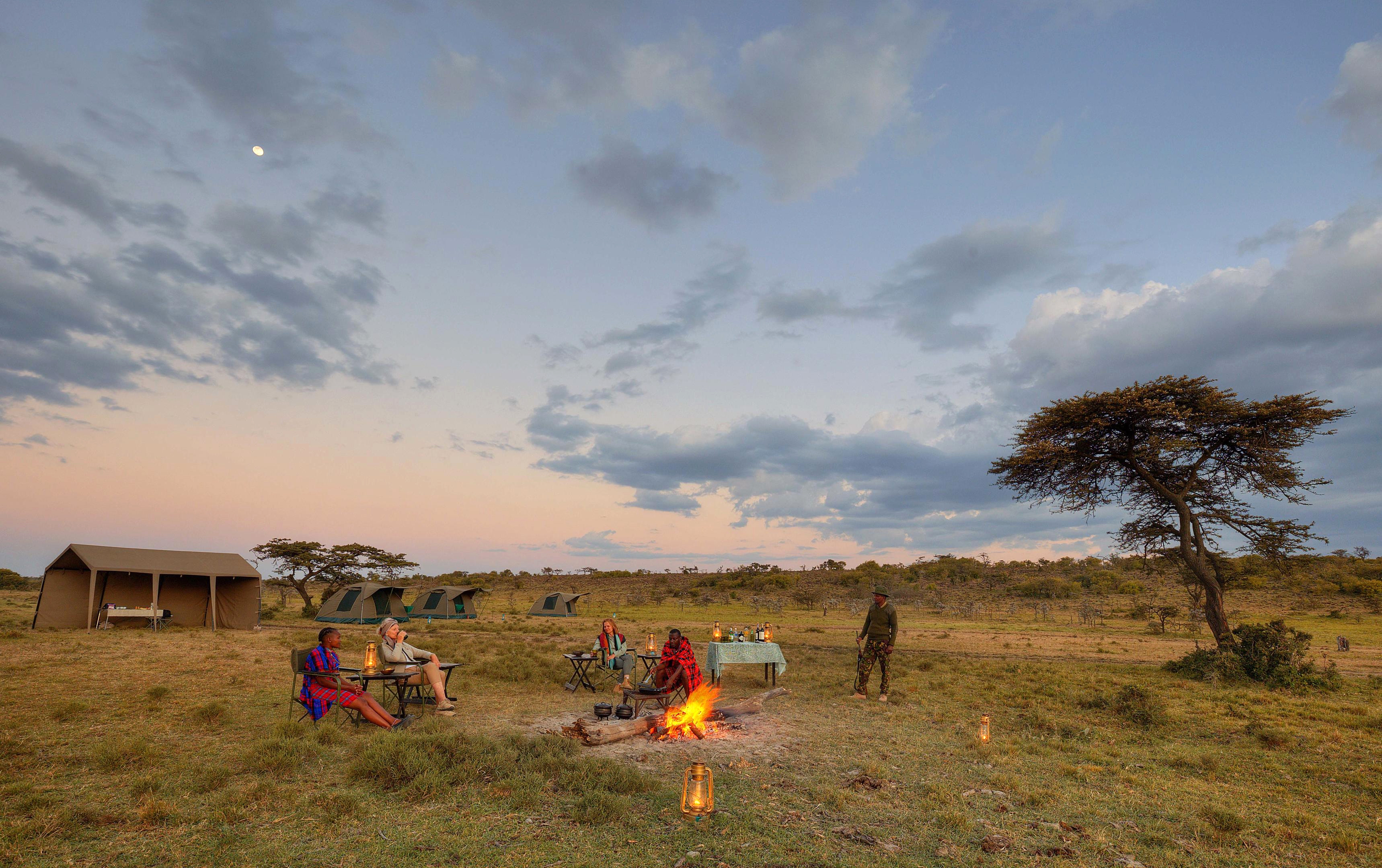 Guests sit around a campfire outside safari tents at dusk, with lantern light and open grassland all around.