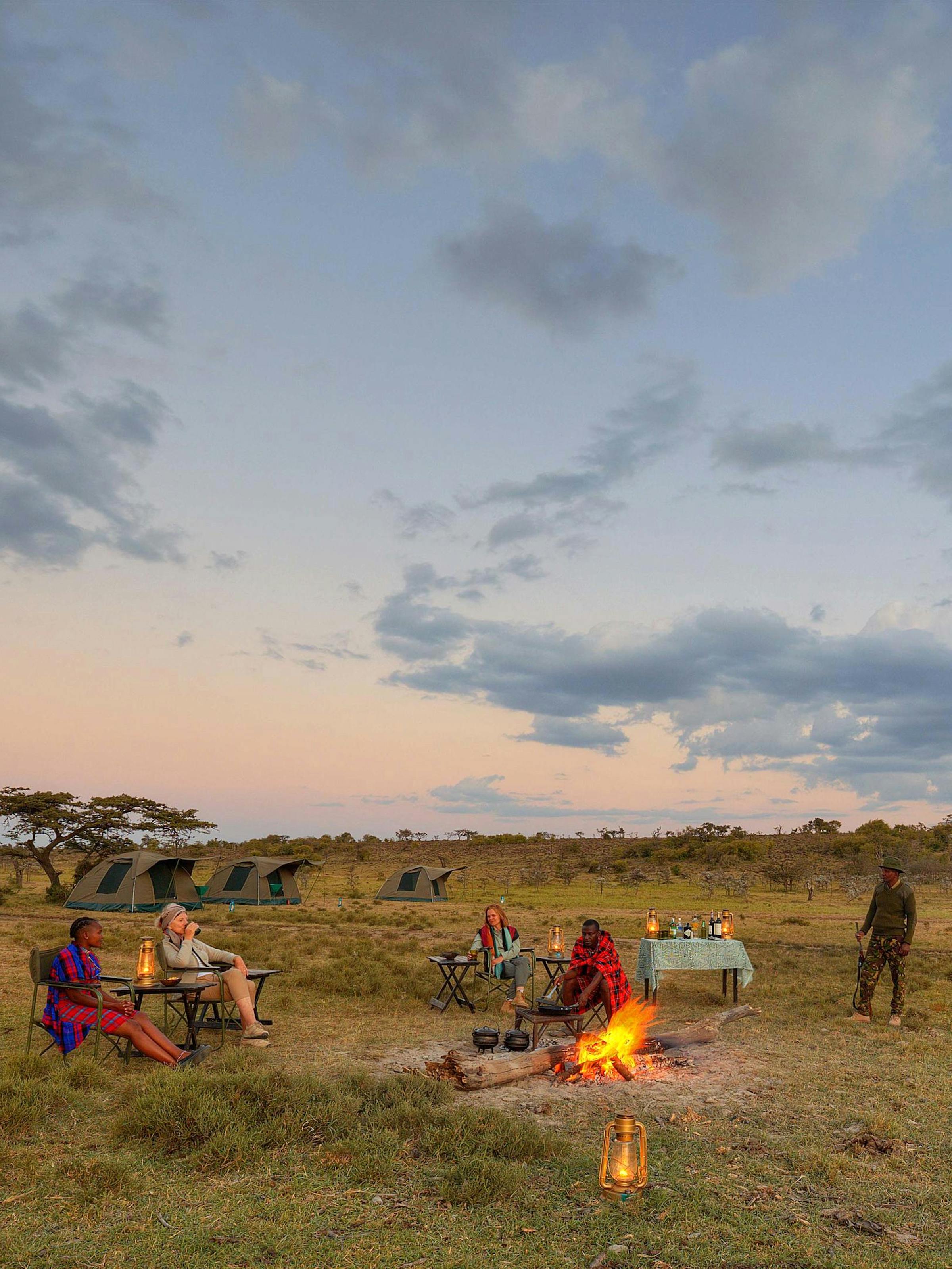 Guests sit around a campfire outside safari tents at dusk, with lantern light and open grassland all around.