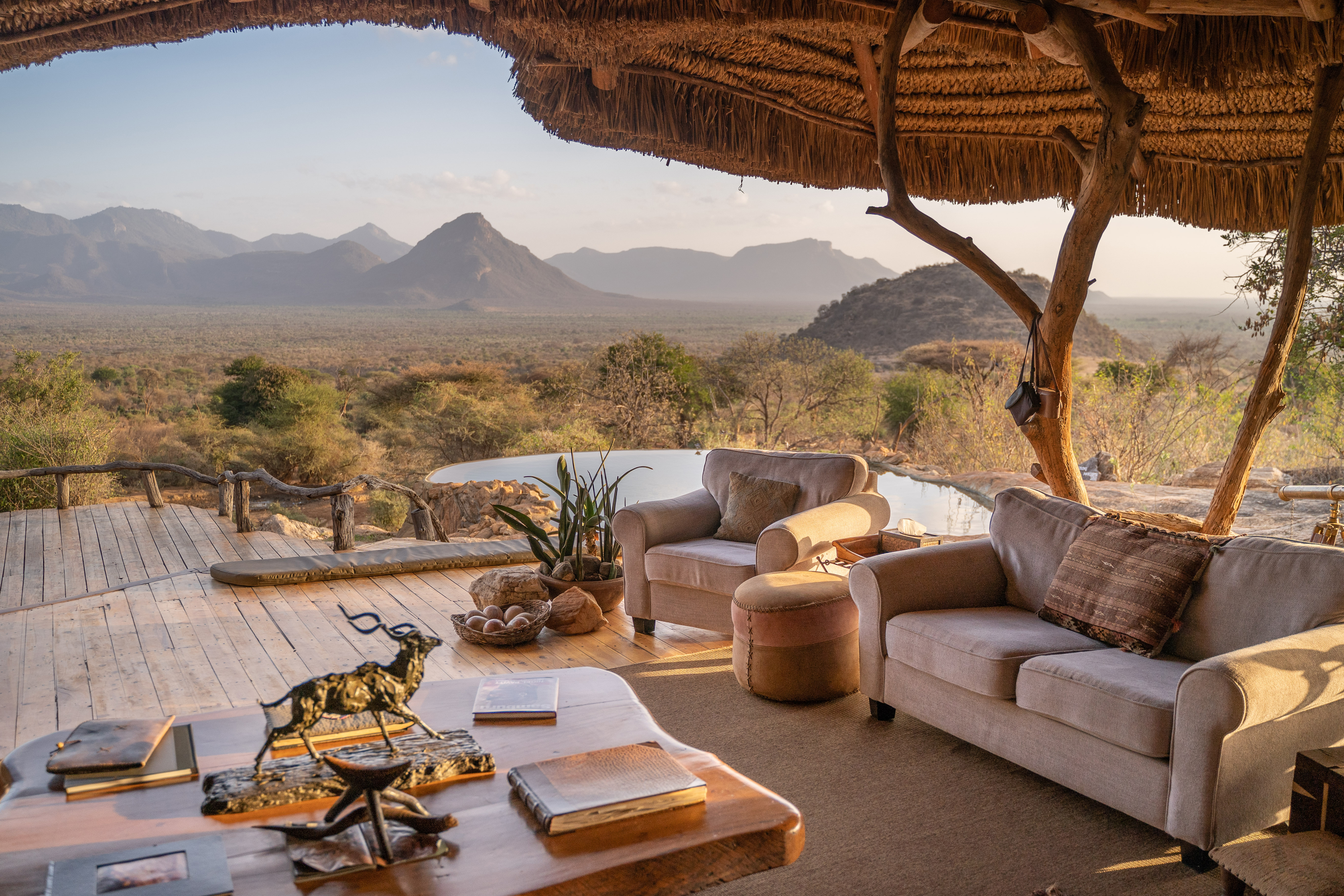 A shaded camp terrace with sofas and a table overlooks a rugged mountain landscape in soft morning light.