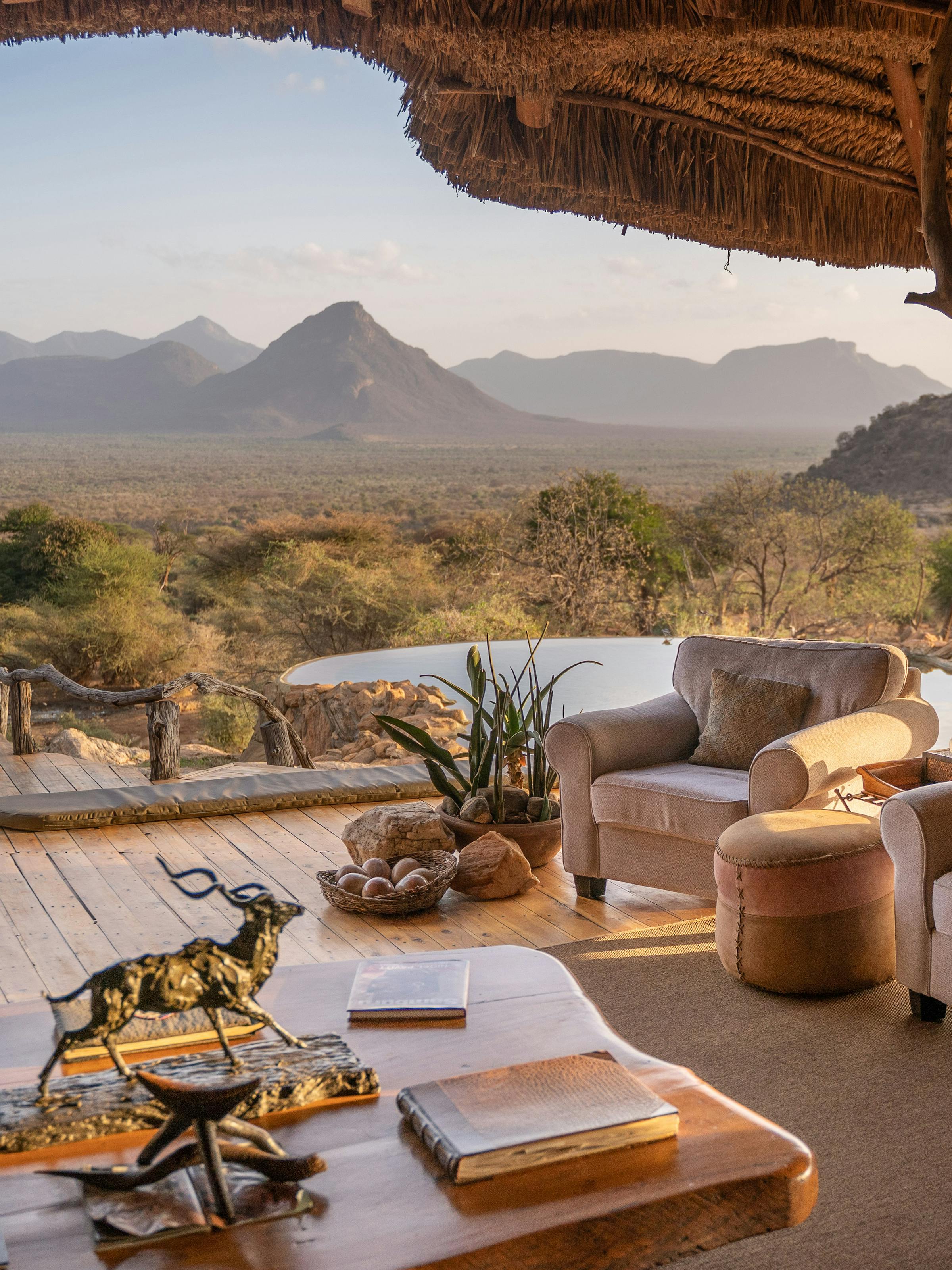 A shaded camp terrace with sofas and a table overlooks a rugged mountain landscape in soft morning light.