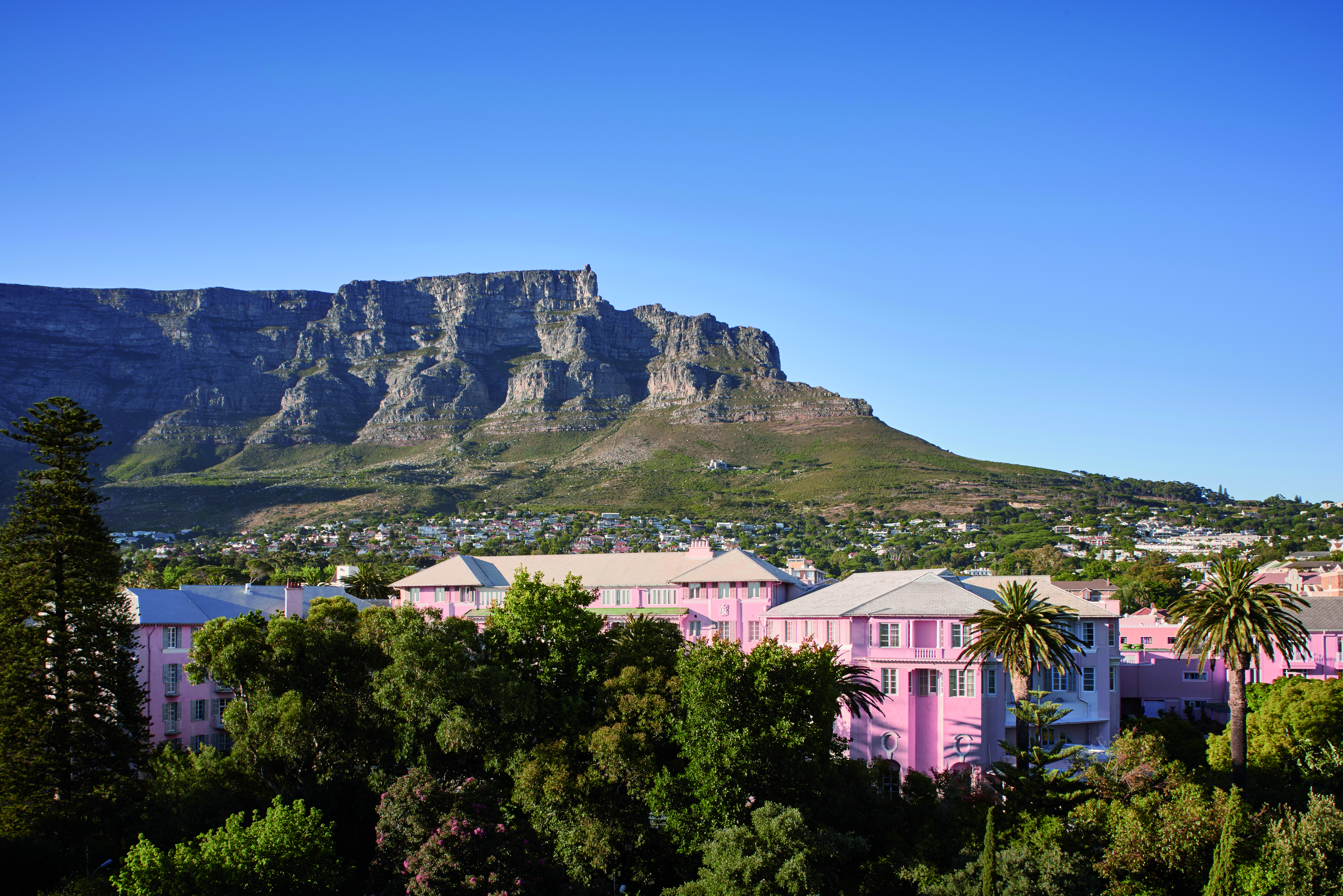 A pink building sits among trees with Table Mountain rising above a sunlit city under a clear blue sky.