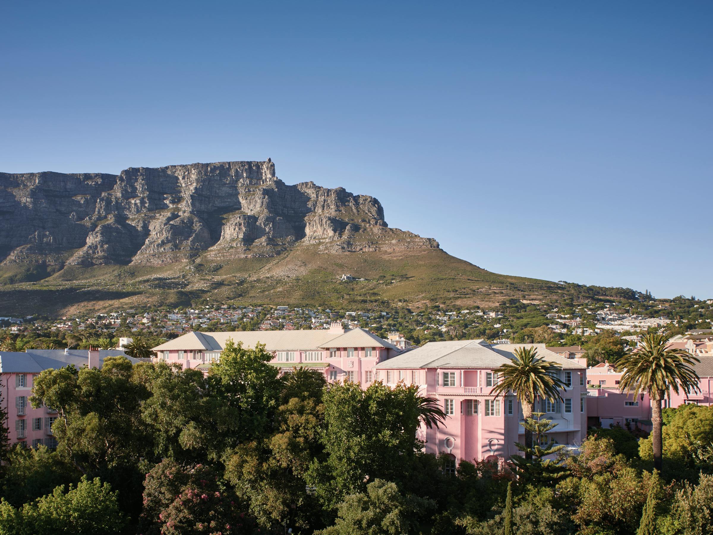 A pink building sits among trees with Table Mountain rising above a sunlit city under a clear blue sky.