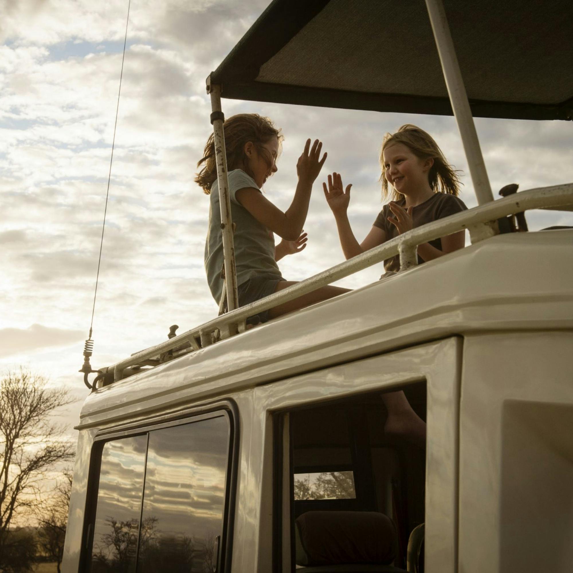 Two travelers stand on a safari vehicle roof at sunrise, looking over the plains as light breaks through clouds.