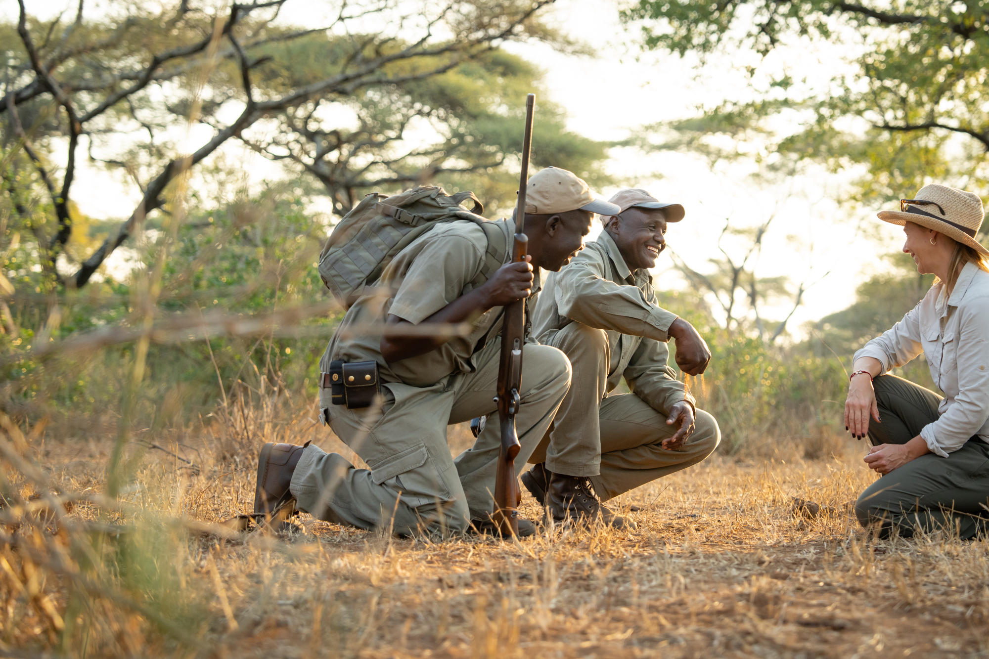 Three safari guides crouch in dry grass beside an open vehicle, studying tracks in warm golden evening light.