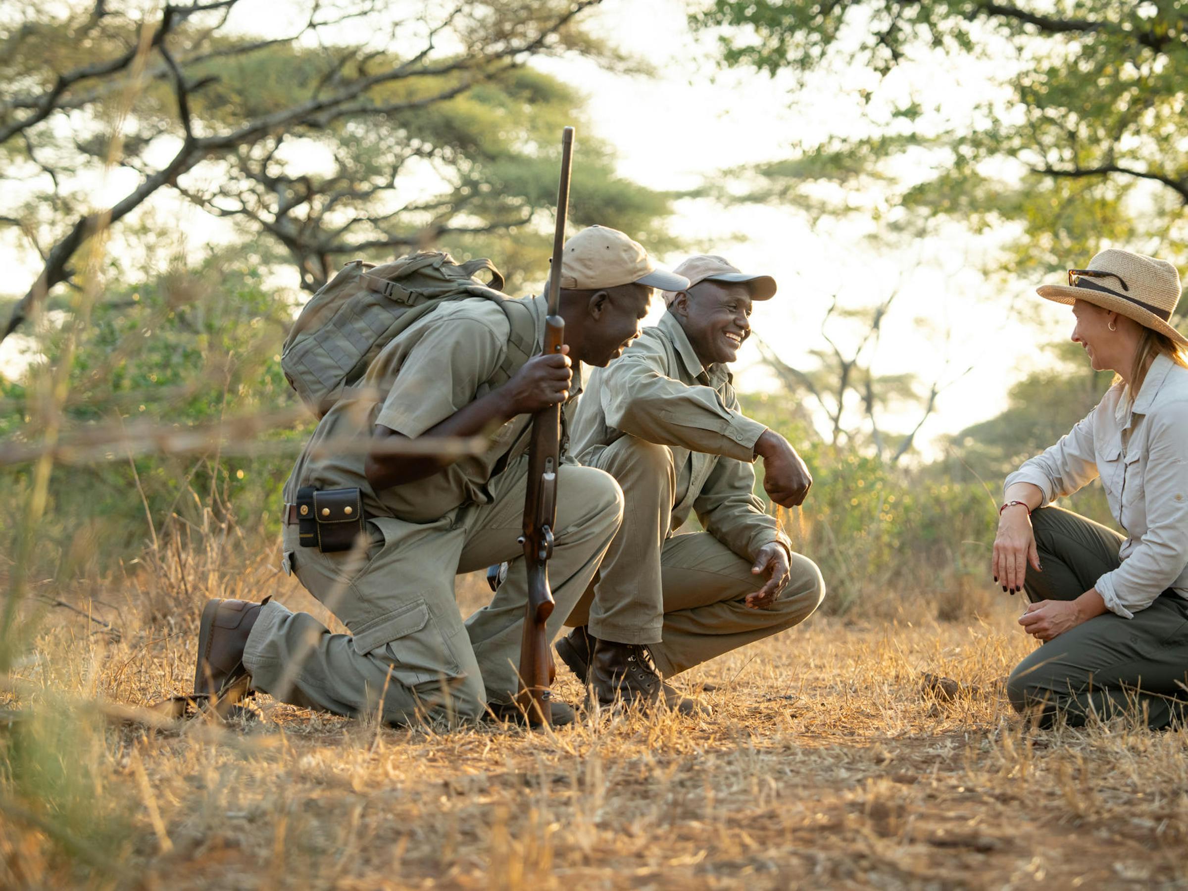 Three safari guides crouch in dry grass beside an open vehicle, studying tracks in warm golden evening light.