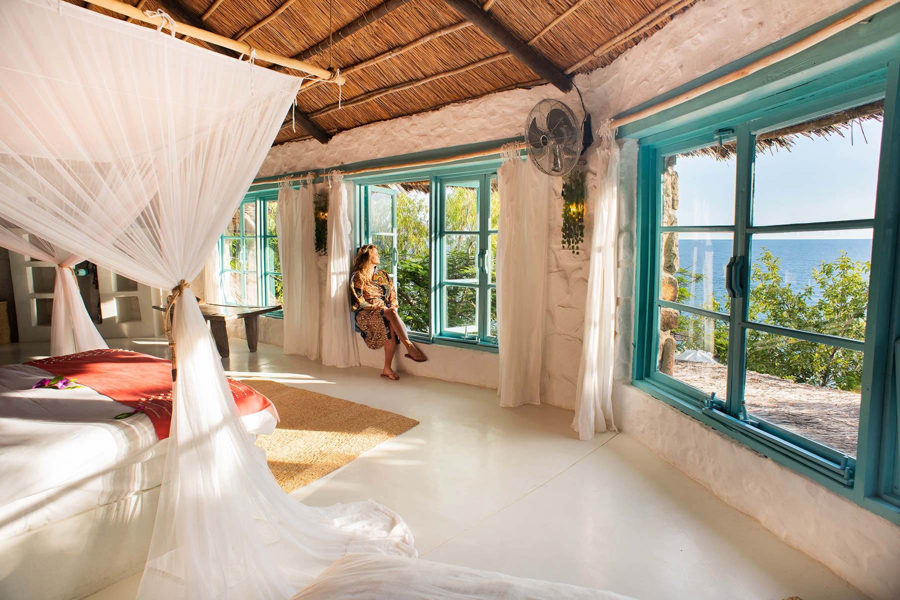Sunlit open-air lodge room with a white bed and mosquito net, wide windows, and a bright blue lake view beyond.