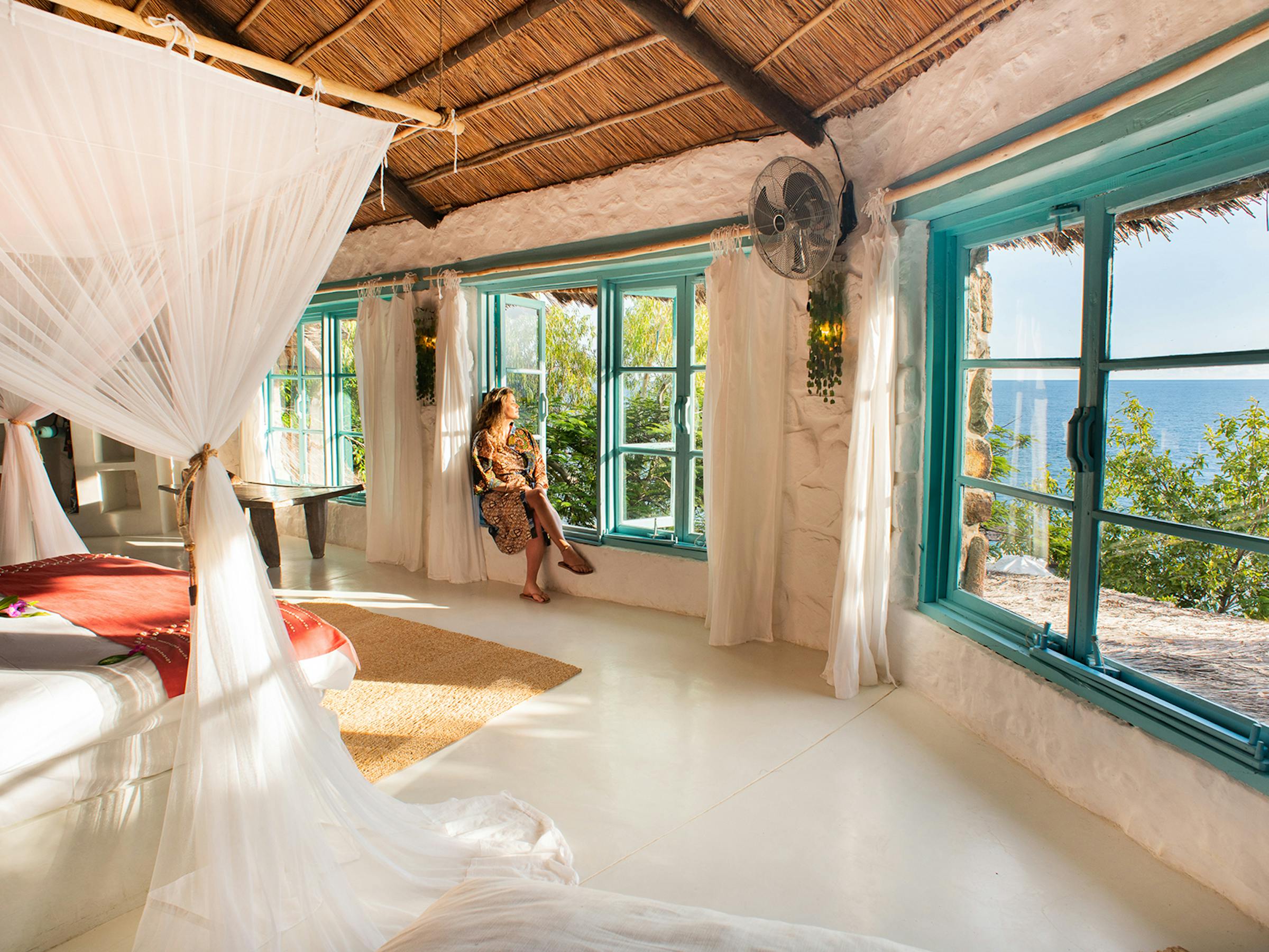 Sunlit open-air lodge room with a white bed and mosquito net, wide windows, and a bright blue lake view beyond.