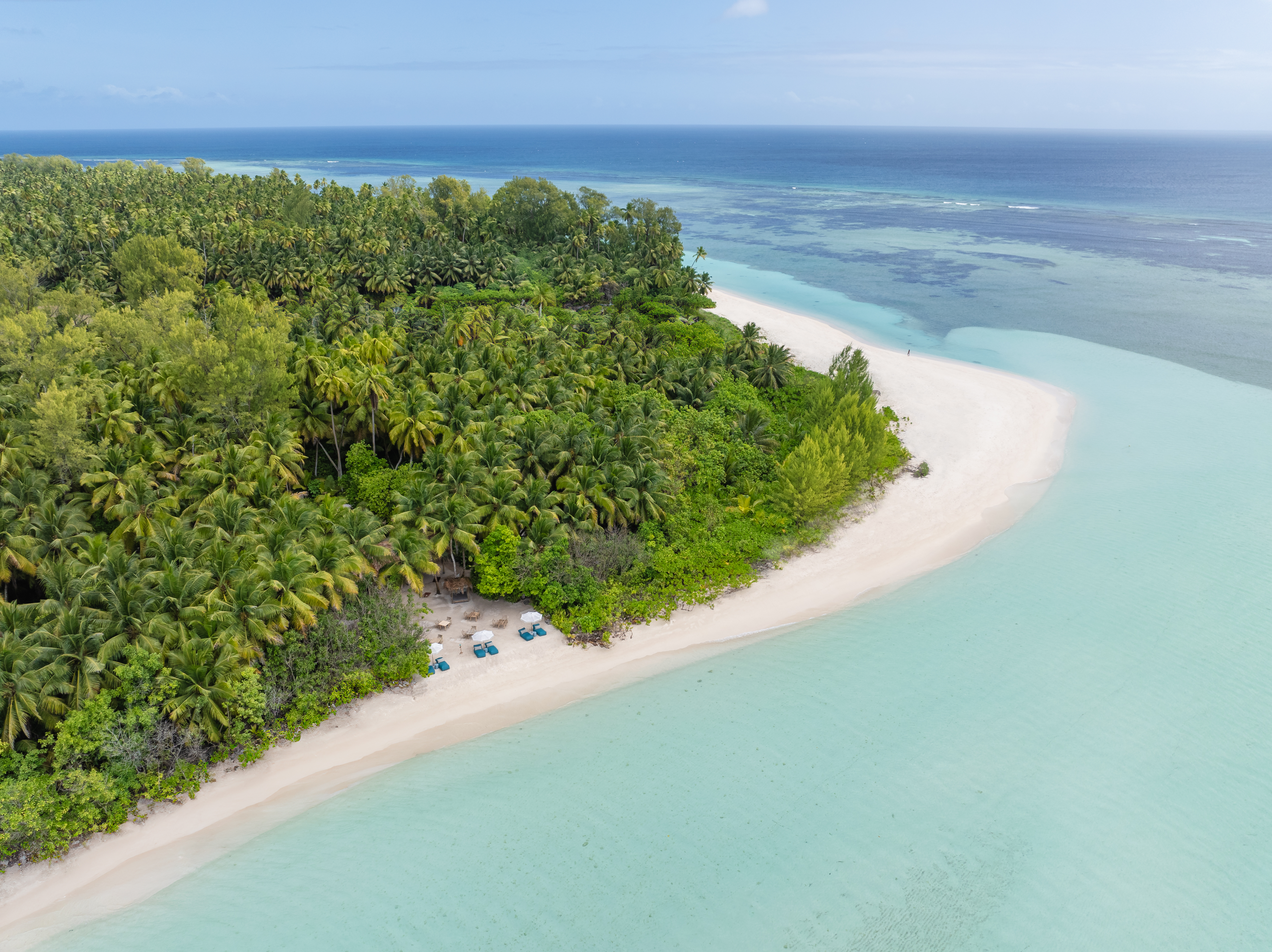 Aerial view of a small tropical island and sandbar, ringed by turquoise lagoon and deeper blue ocean under scattered clouds.