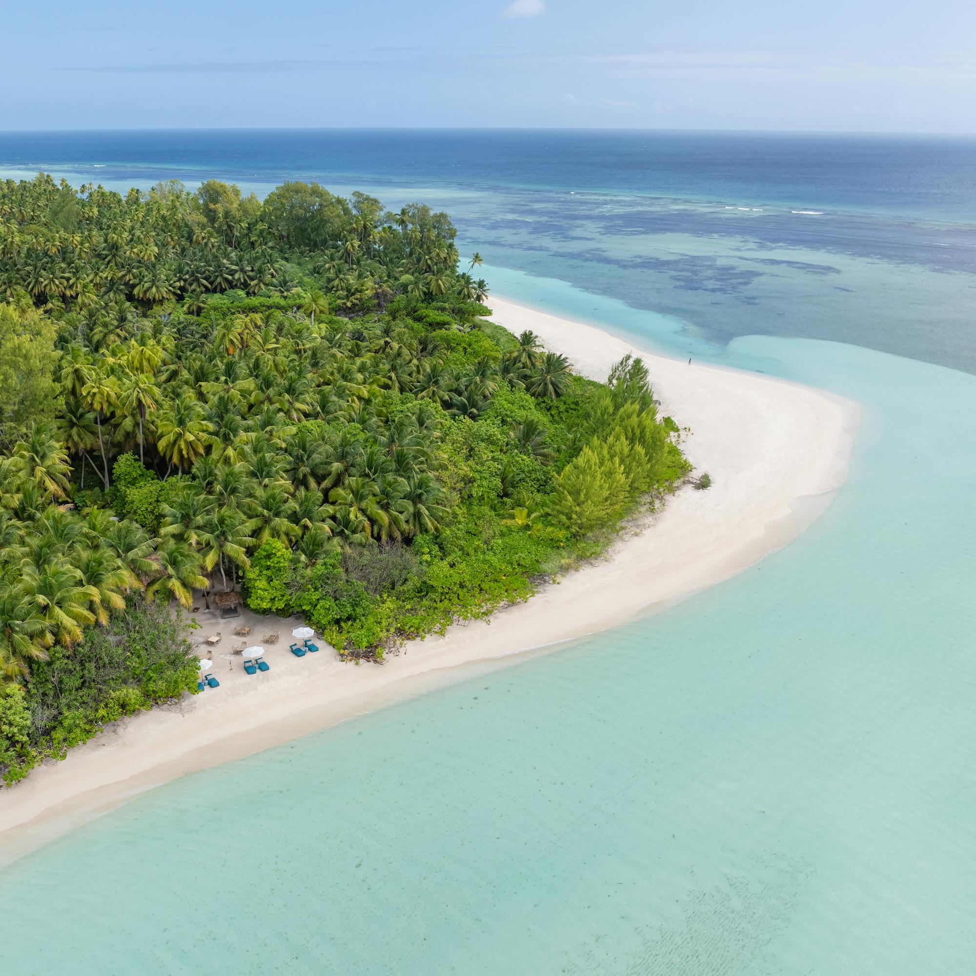 Aerial view of a small tropical island and sandbar, ringed by turquoise lagoon and deeper blue ocean under scattered clouds.