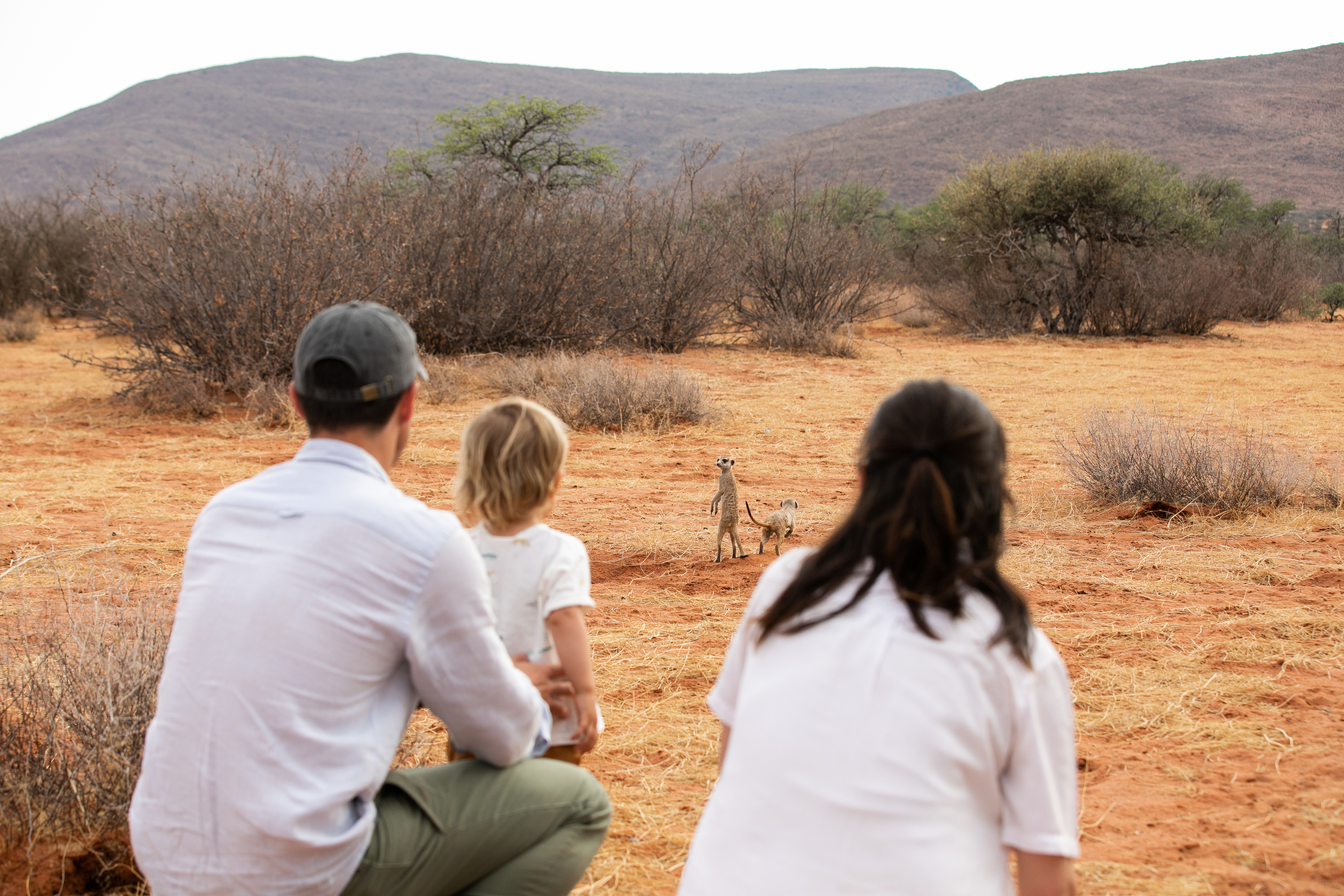 Three people sit on a sandy rise, watching distant hills and open plains in the warm light of late day together.