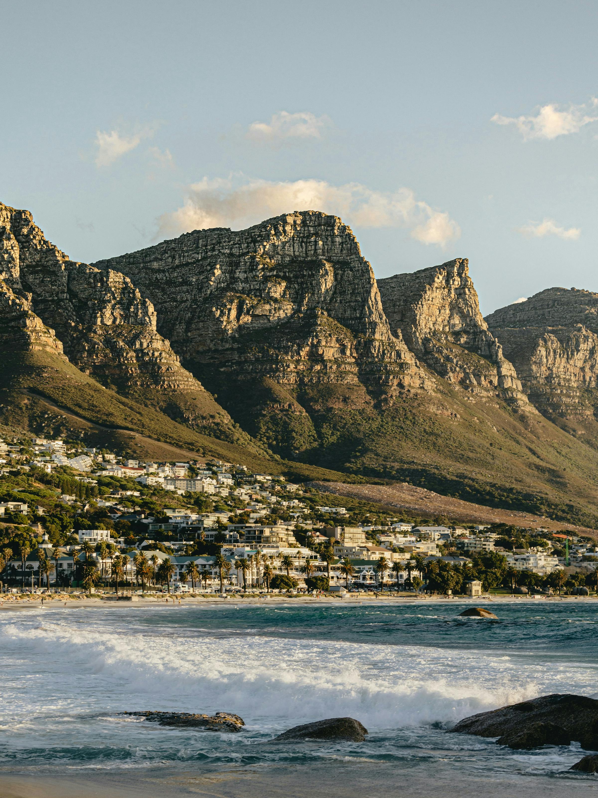 Coastal city spreads along a blue bay below a flat-topped mountain, with boats and buildings in view under a clear sky.