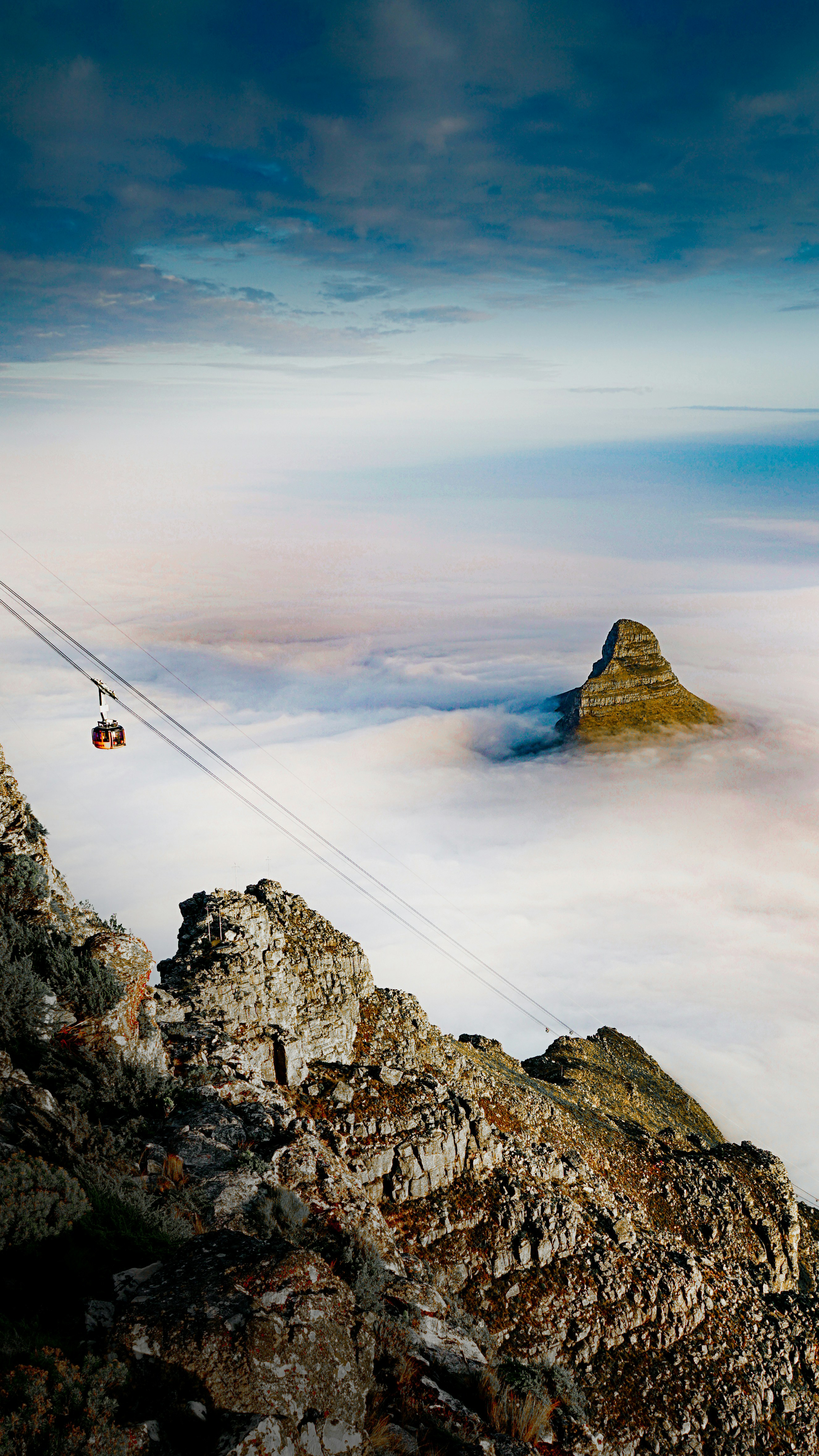 A cable car crosses rocky cliffs above a blanket of clouds, with a steep peak rising through the mist.