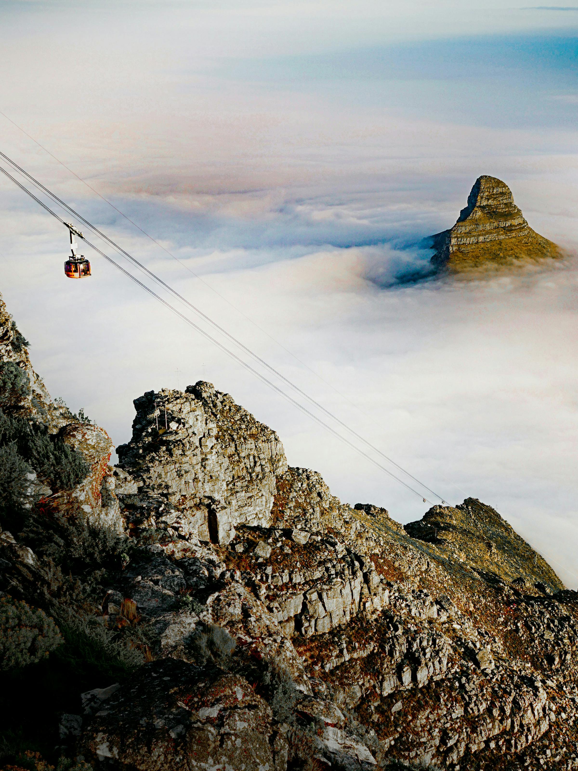 A cable car crosses rocky cliffs above a blanket of clouds, with a steep peak rising through the mist.