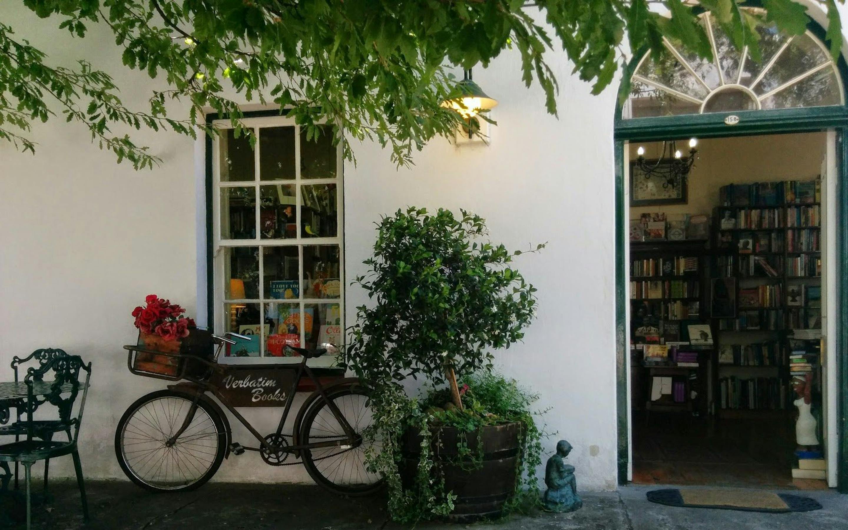 A bicycle leans beside a café doorway framed by vines and potted plants, with chairs set on a stone patio.